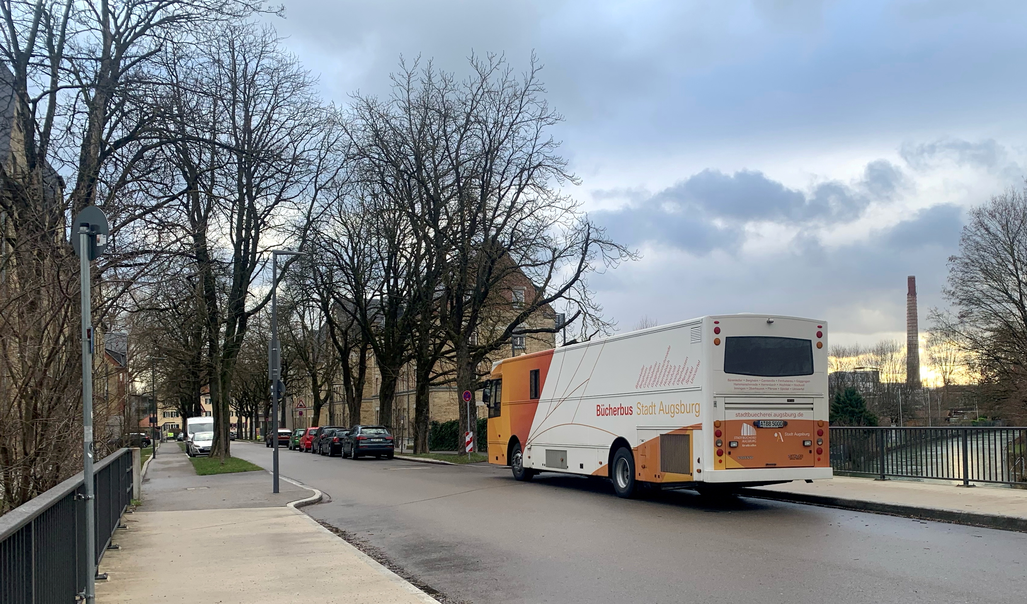 ein Bus in orange und weiß und der Aufschrift "Bücherbus Stadt Augsburg" fährt auf einer leeren Straße. 