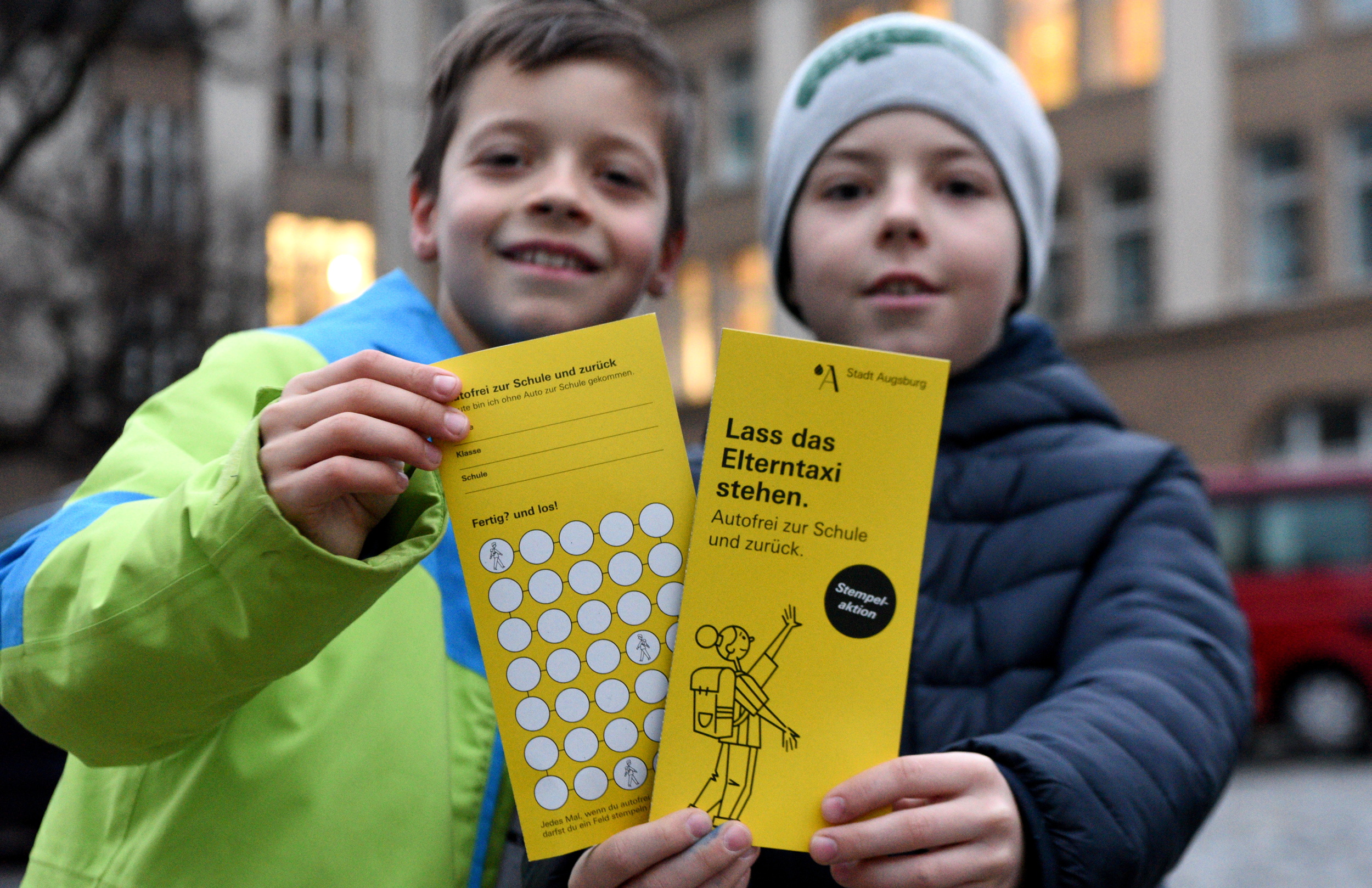 zwei Kinder halten gelbe Stempelkarten mit der Aufschrift "Lass das Elterntaxi Stehen" in die Kamera.