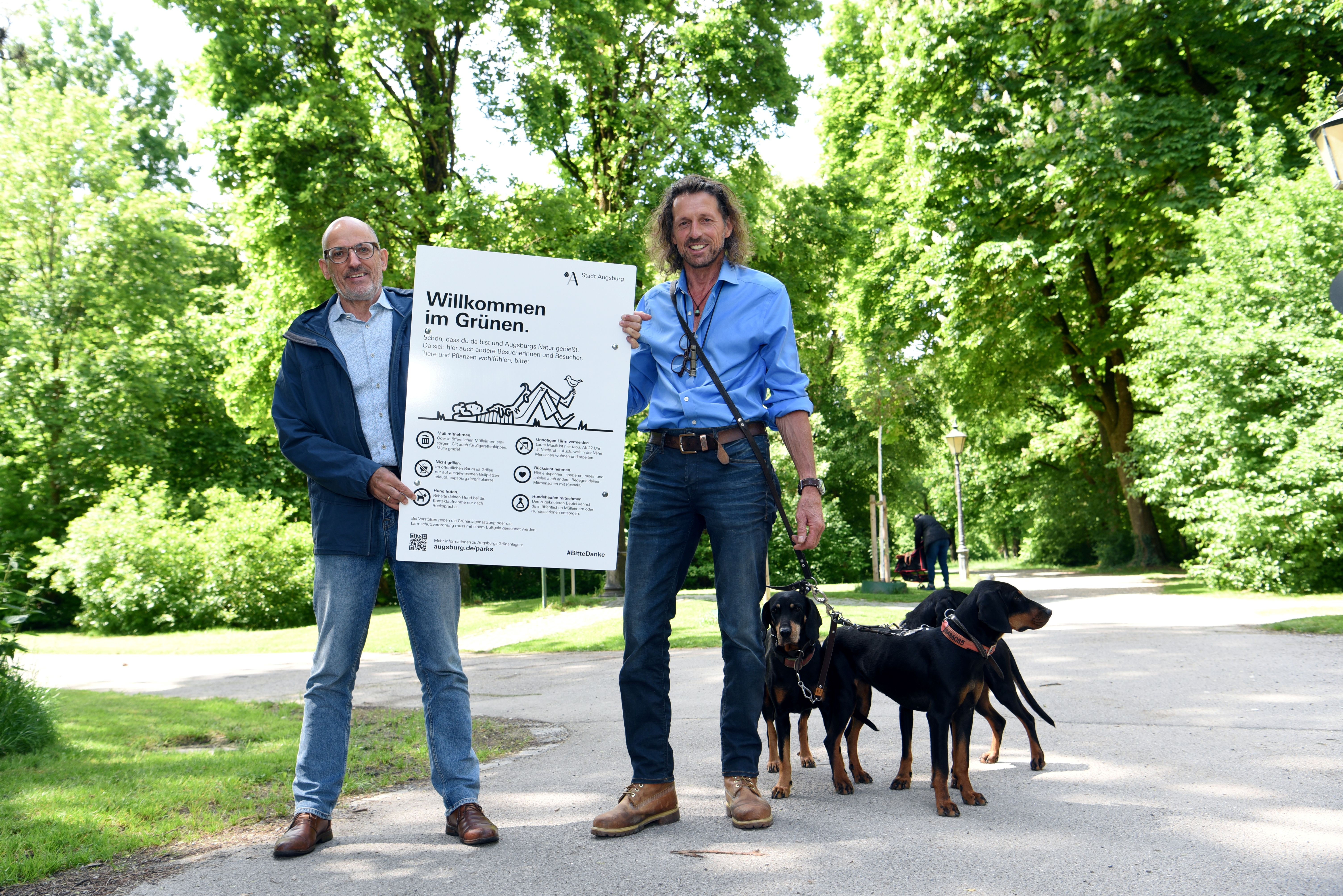 Zwei Männer mit Hunden stehen unter Bäumen und halten ein Schild in der Hand