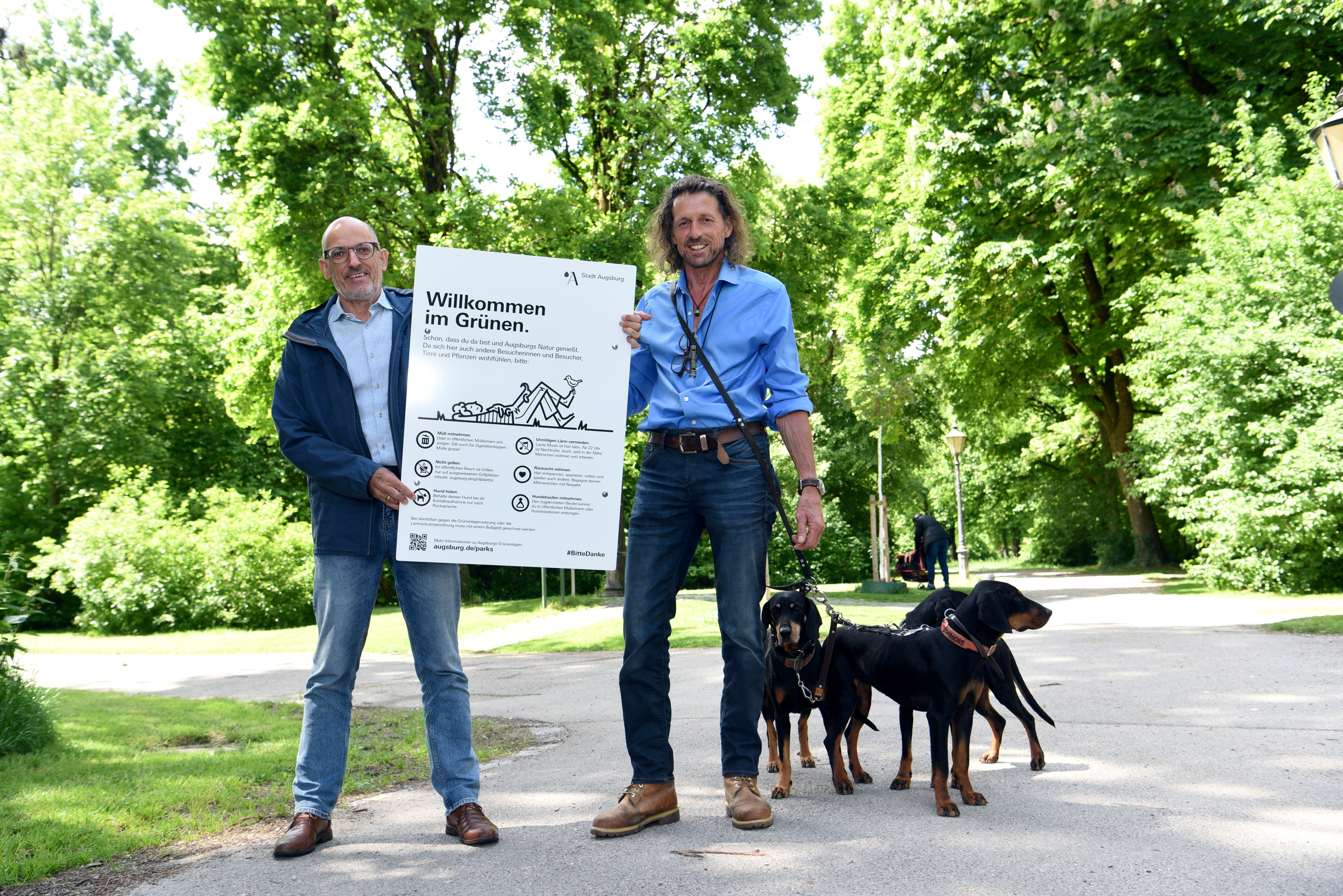 Zwei Männer mit Hunden stehen unter Bäumen und halten ein Schild in der Hand