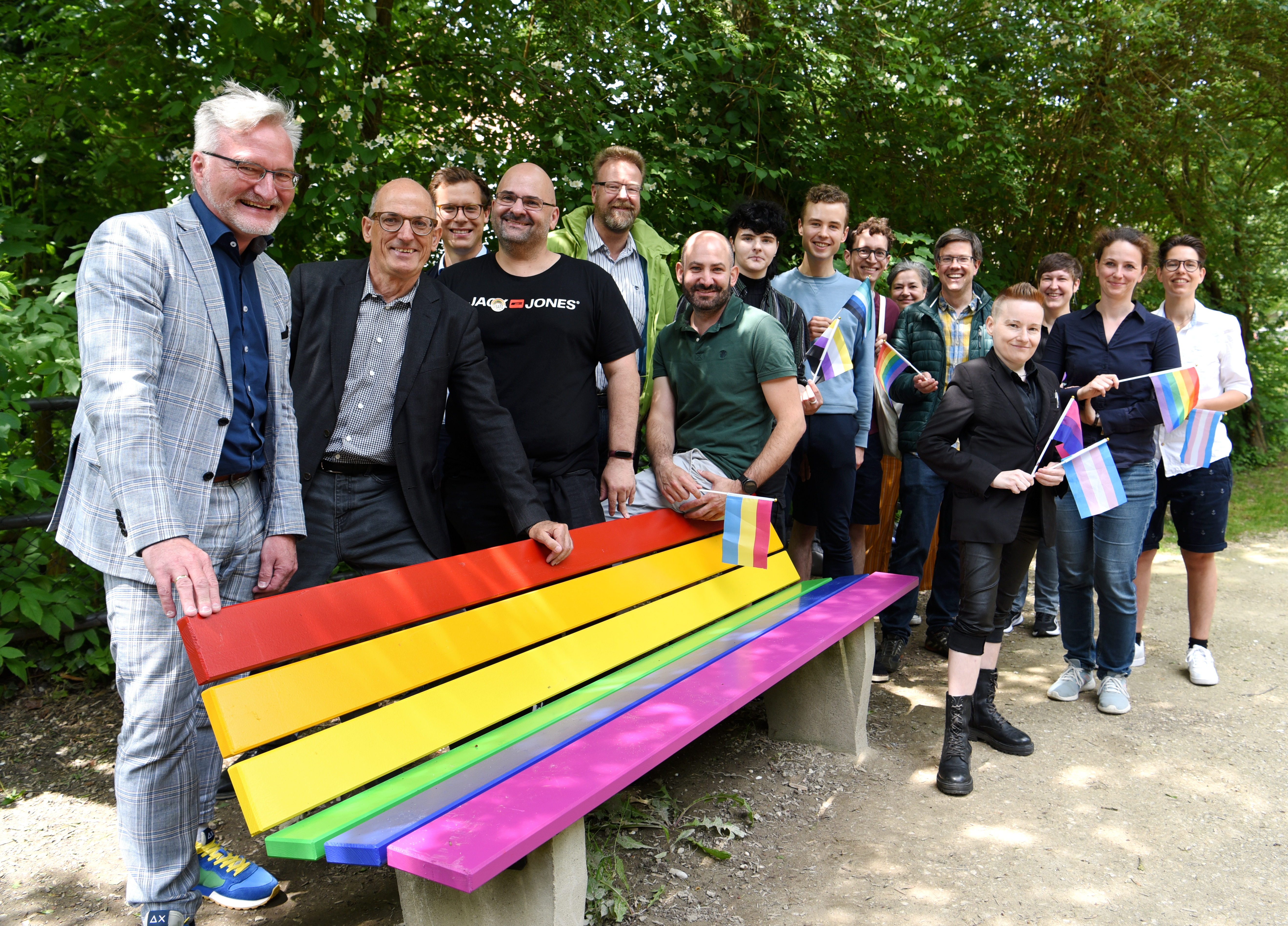 Etwa 15 Leute stehen hinter einer Parkbank, die in bunten Farben angemalt ist.