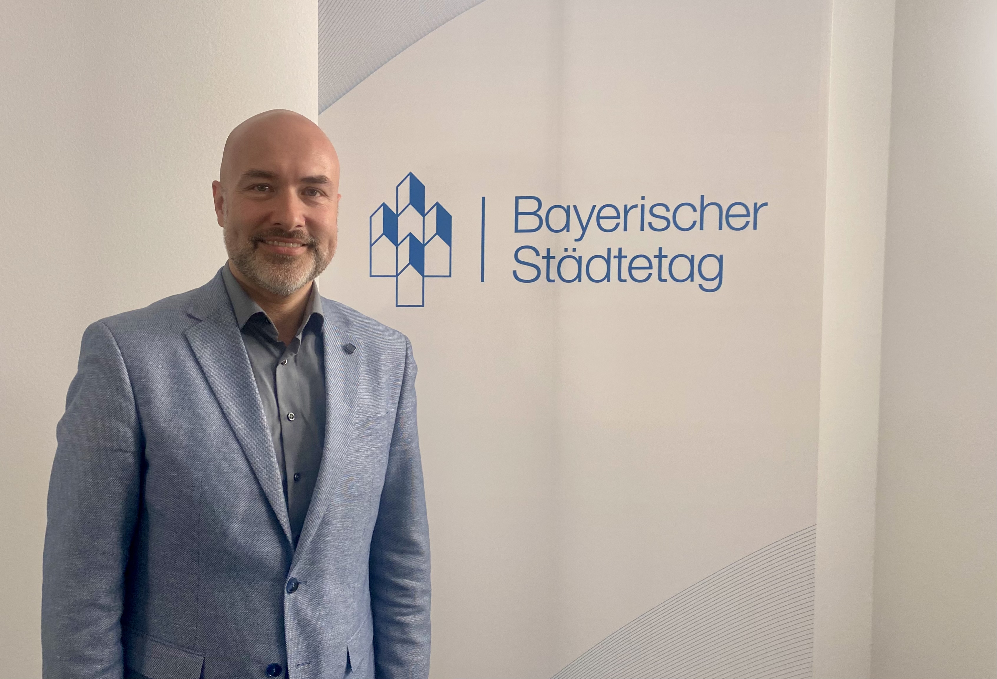 Martin Schenkelberg steht in einem grauen Anzug vor einem Plakat mit der Aufschriift "Bayerischer Städtetag"