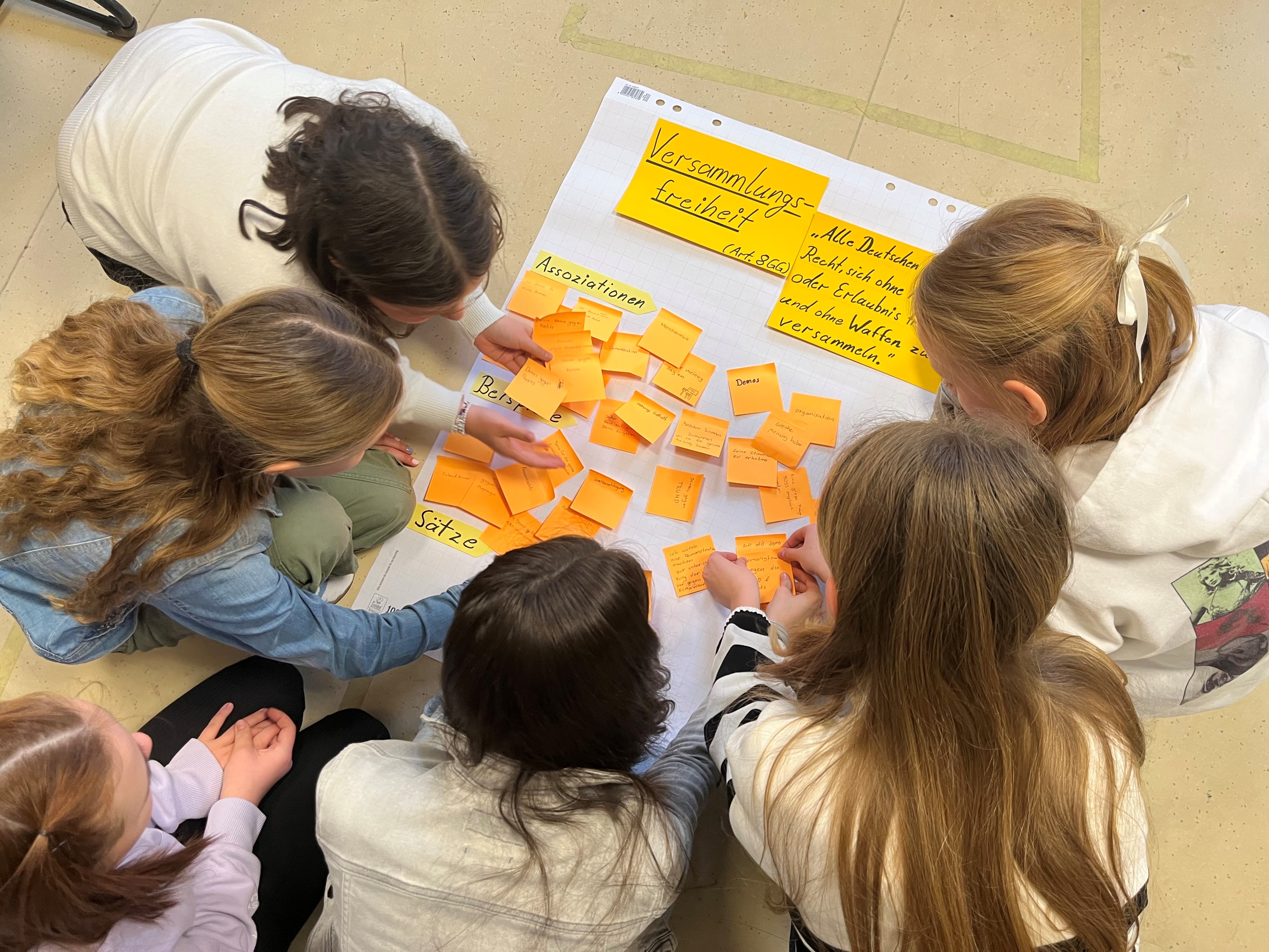 Blick von oben auf eine Gruppe Kinder, die um ein auf dem Fußboden liegendes Plakat mit dem Titel "Versammlungsfreiheit" herumsitzen.