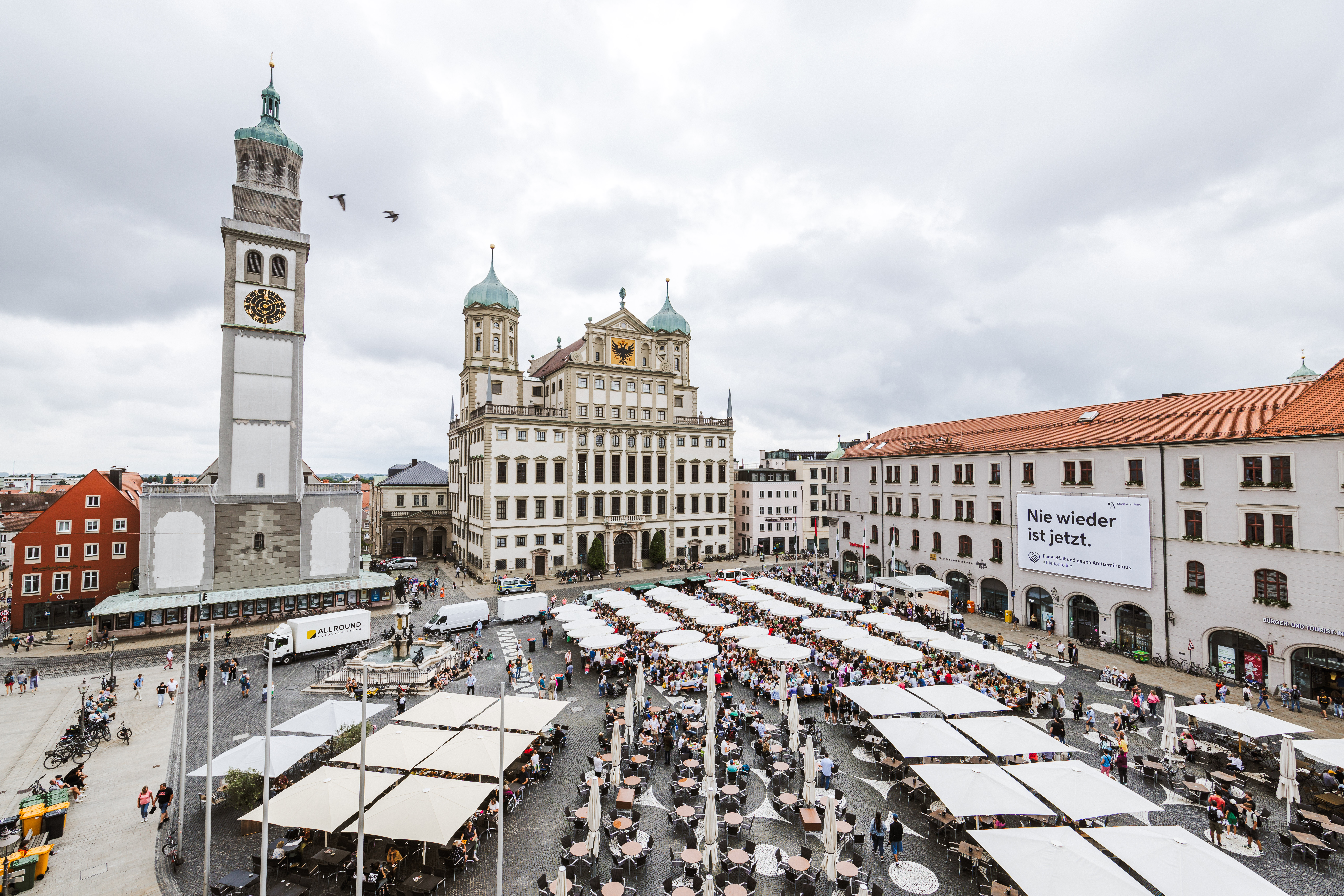 Auf dem Rathausplatz sind viele Tische mit Sonnenschirmen aufgebaut. Viele Menschen sitzen dort und essen