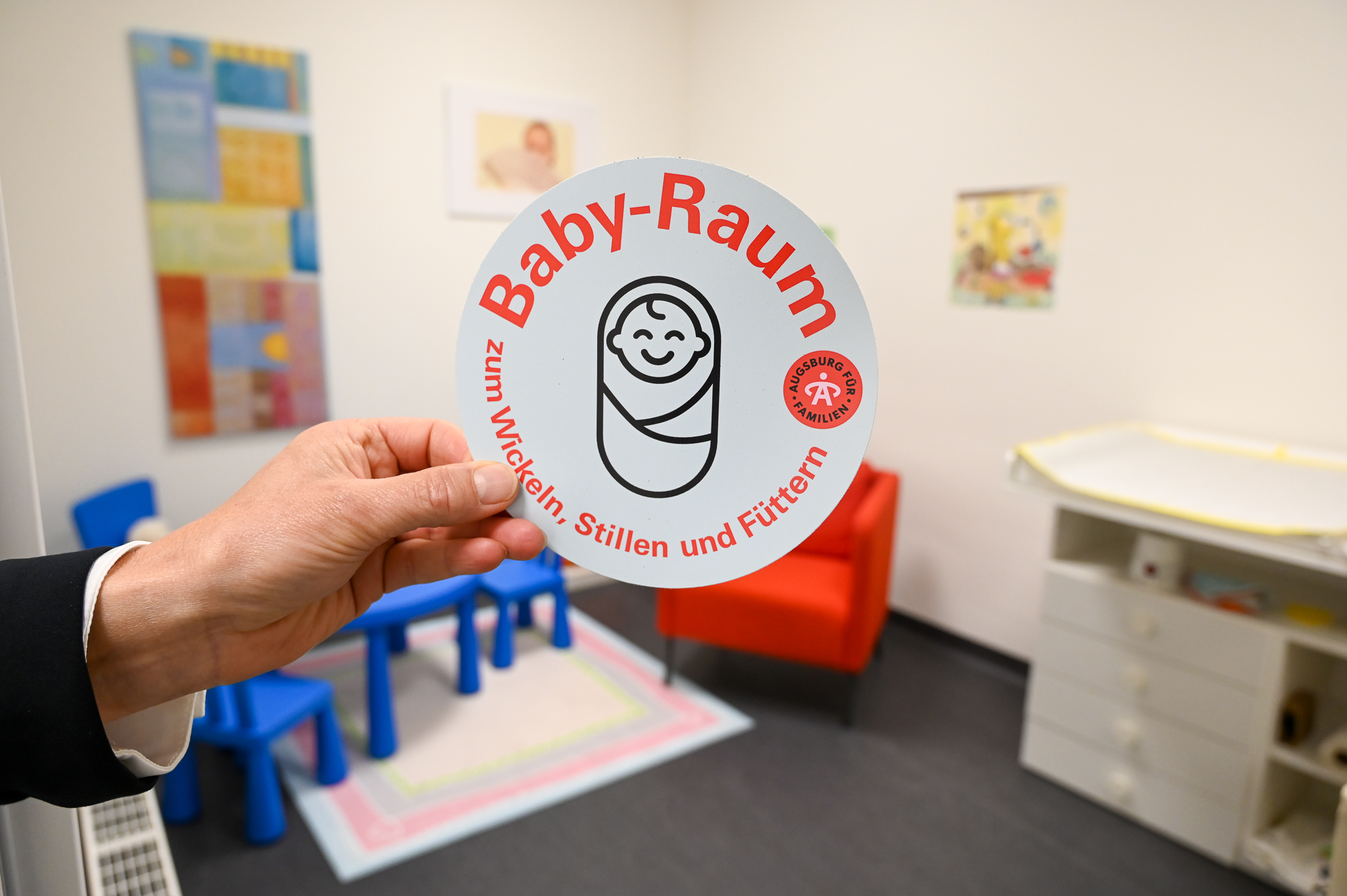 Der Hinweis-Aufkleber Baby-Raum wird von einer Hand gehalten. Im Hintergrund ist ein Beispielraum zu sehen.