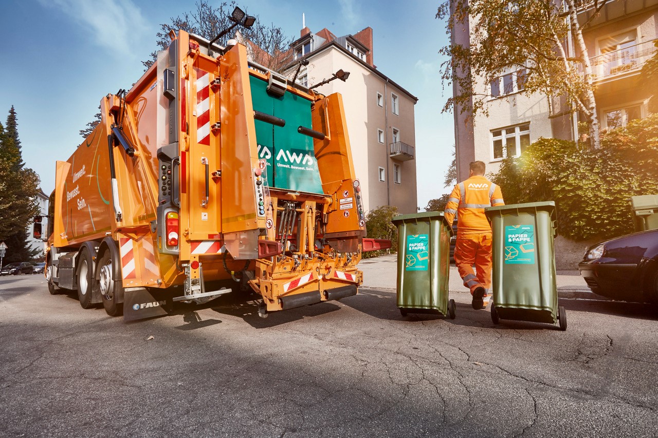 Großes Müllauto. Ein Mitarbeiter fährt zwei Altpapier-Tonnen zurück in ein Grundstück.