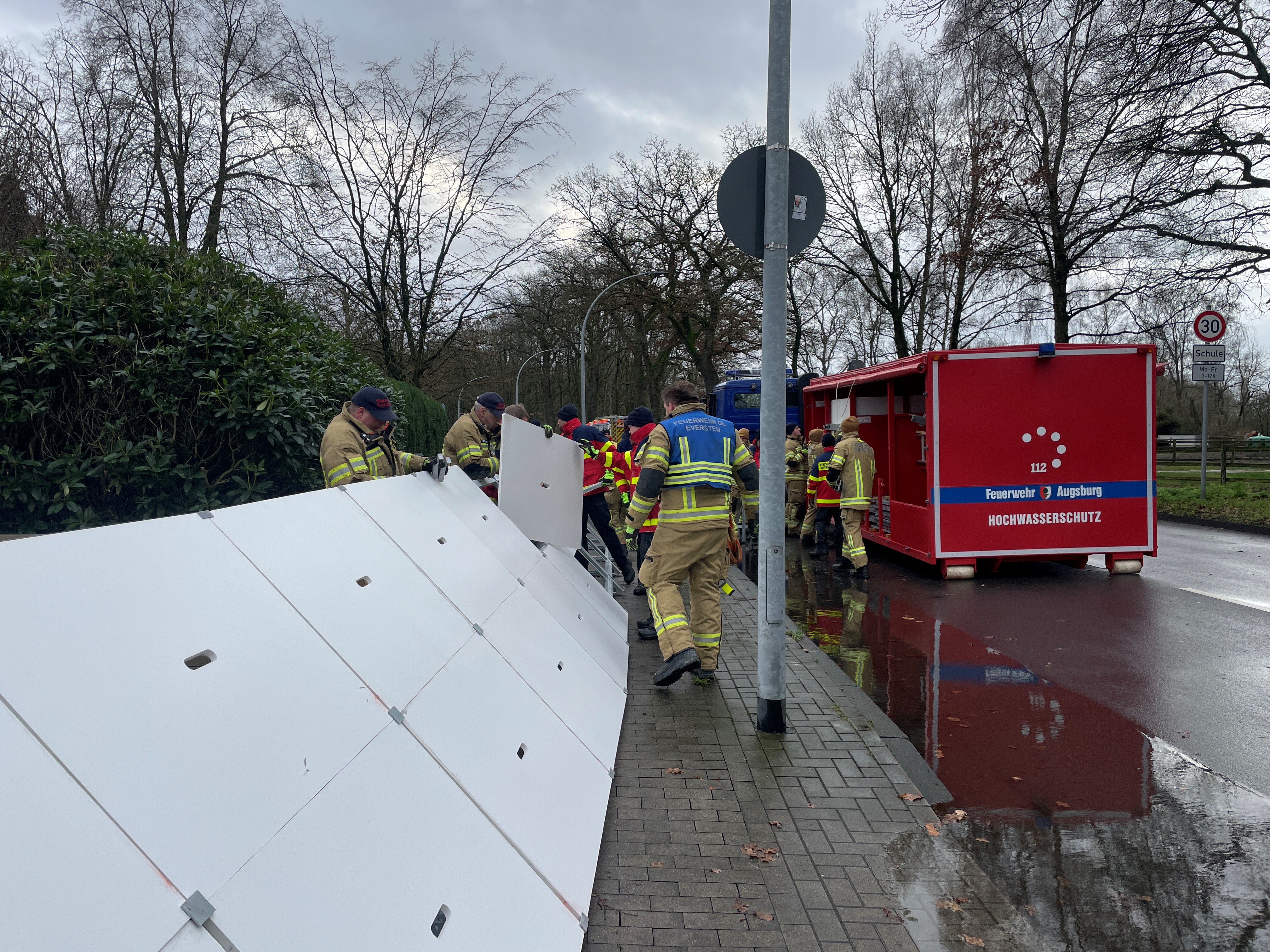 Feuerwehrleute kombinieren Einzelelemente zu einer Schutzwand; auf der Straße steht ein roter Transportcontainer mit der Aufschrift Feuerwehr Augsburg, Hochwasserschutz.