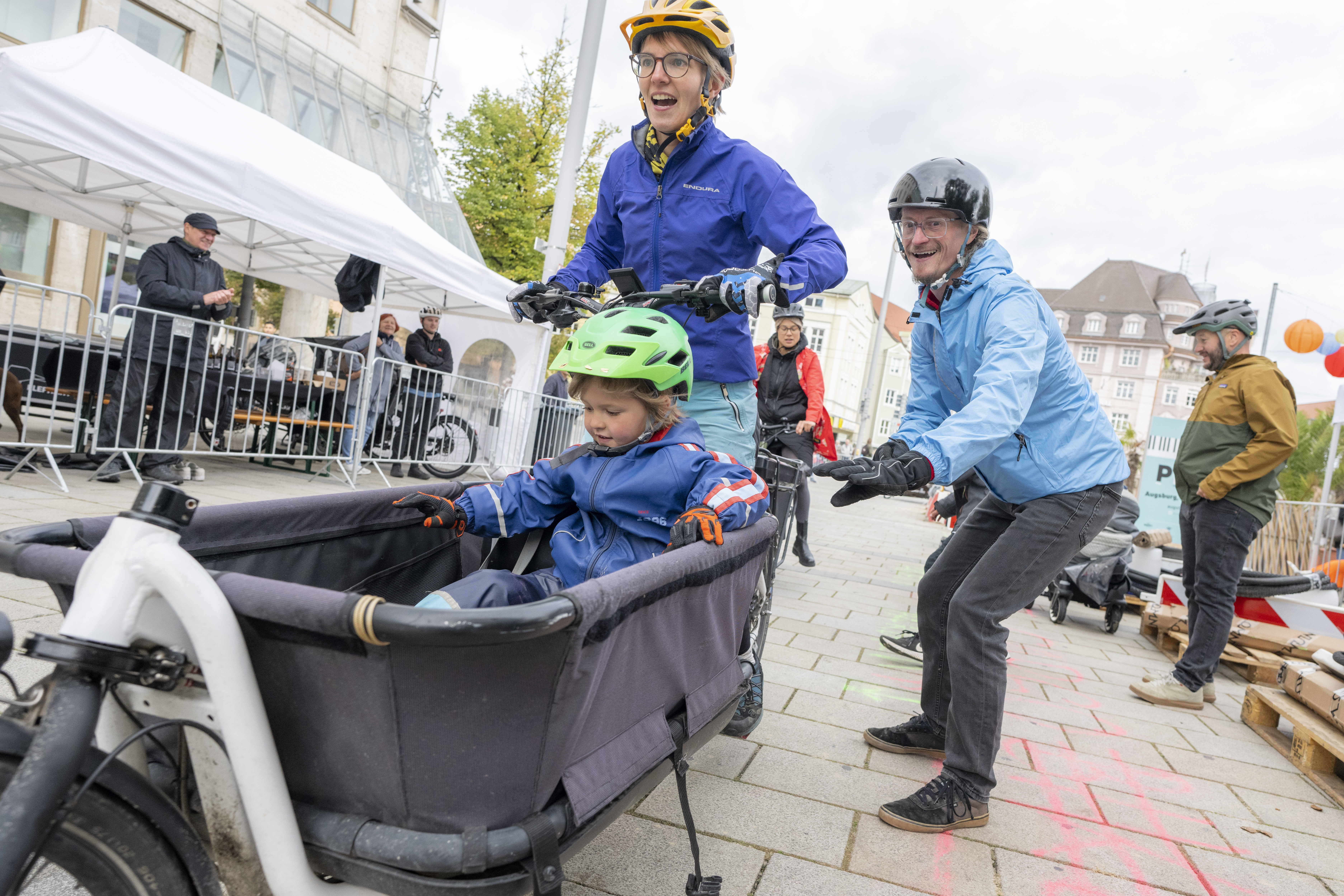 Ein Kind sitzt im Korb eines Lastenrades. Die Mutter sitzt am Lenker. Der Vater steht hinter dem Lastenrad und lacht