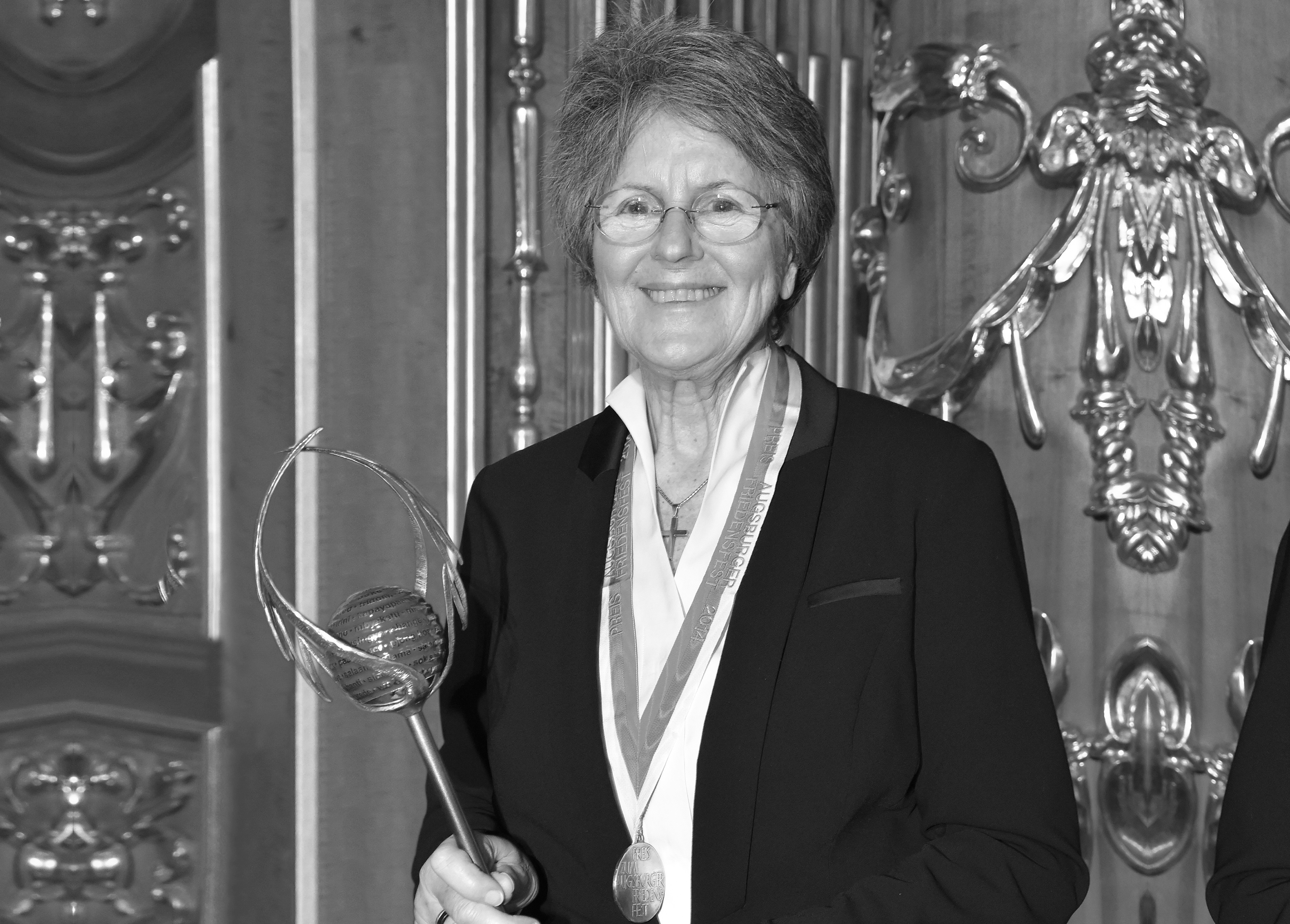 Schwarzweiß-Bild von Dr. Lea Ackermann. Sie hält den Friedenspreis der Stadt Augsburg in der Hand und hat eine Medaille umgehängt. Im Hintergrund sind goldene Ornamente an einer Holzwand zu sehen.