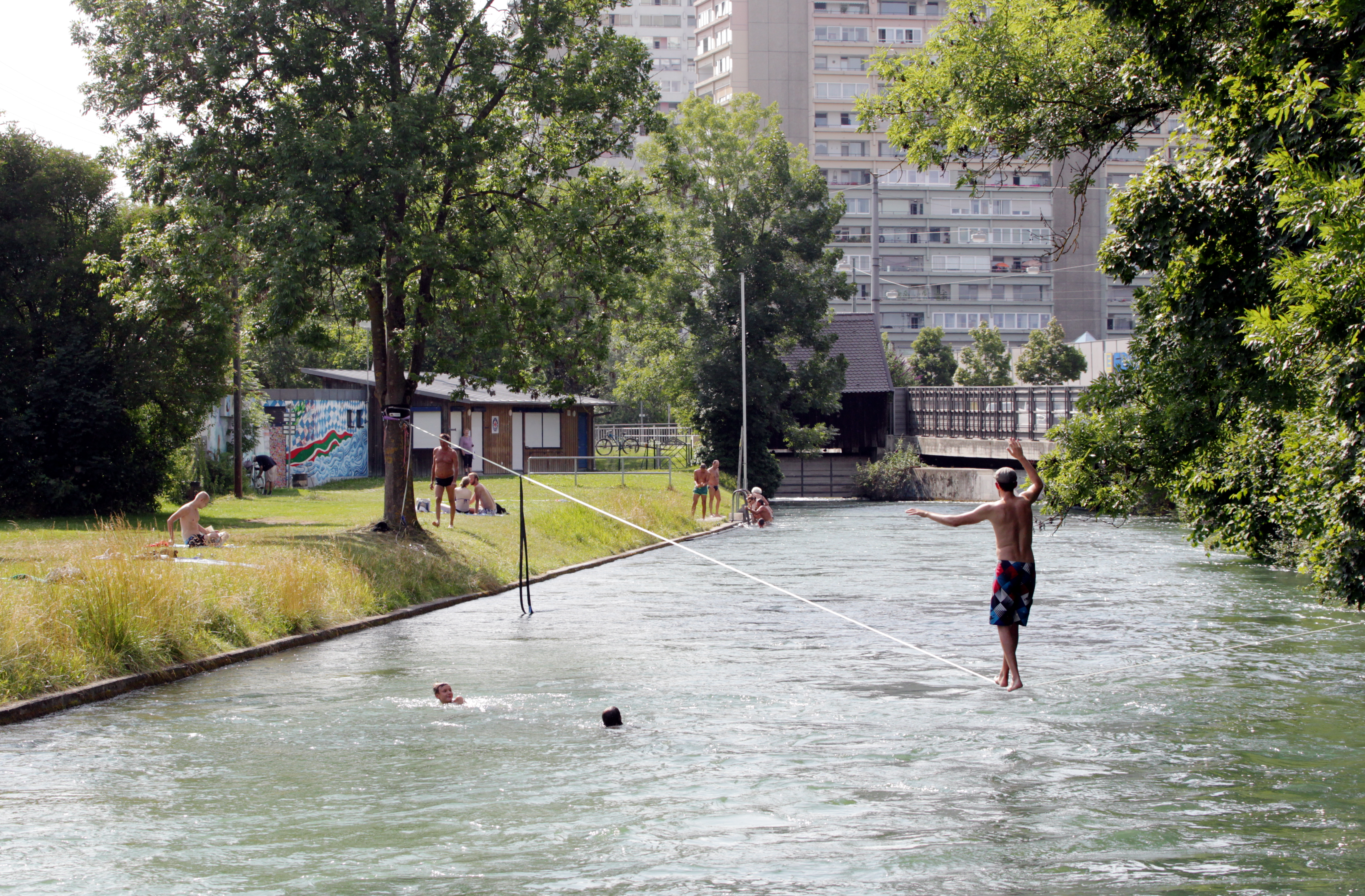 über wasserführenden Kanal balanciert ein junger Mann in Shorts; im Hintergrund ist ein Hochhaus zu sehen