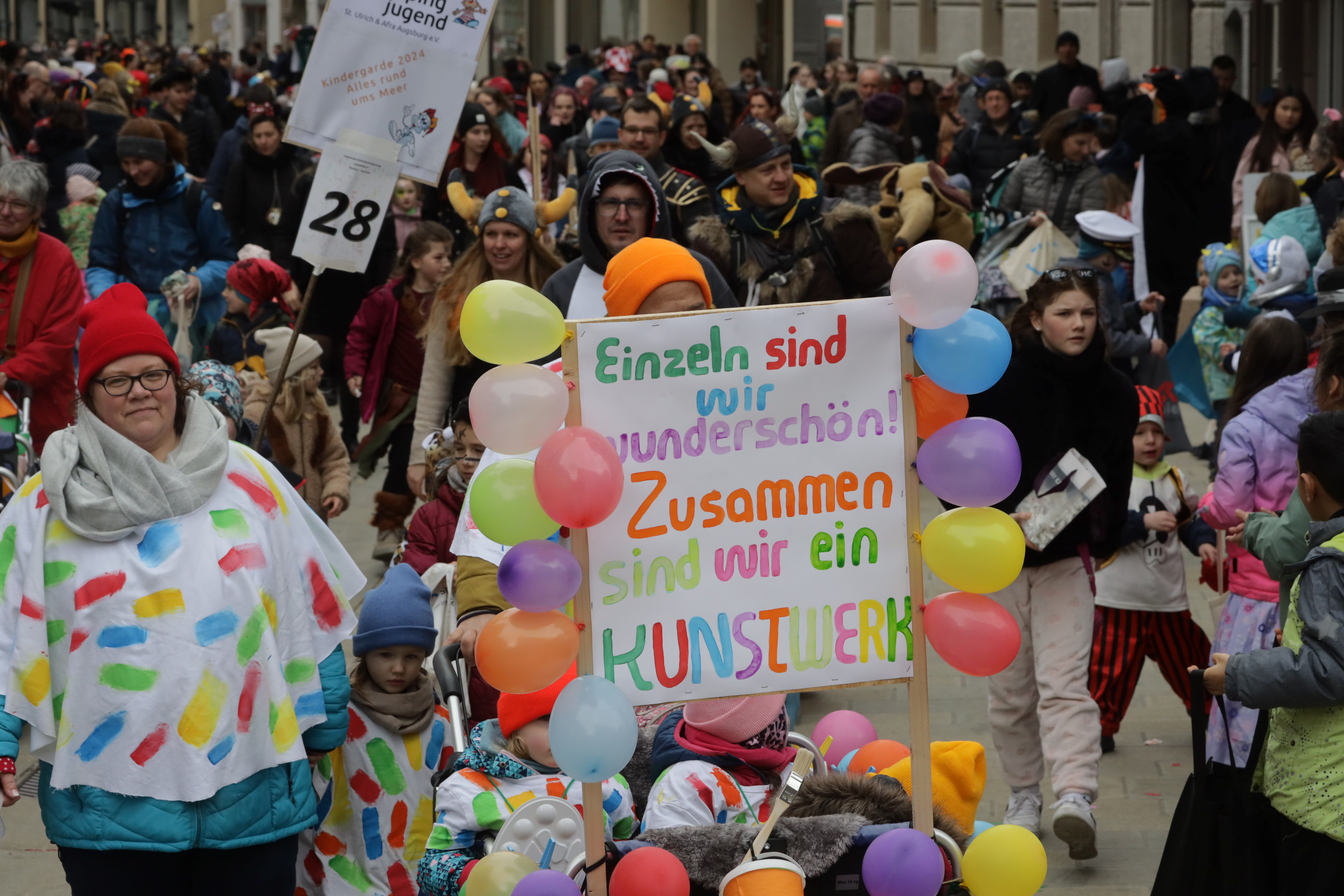 Ein bunter Gruppe an Menschen in Kostümen mit einem Schild in der Hand mit der Aufschrift "Einzeln sind wir wunderschön, zusammen sind wir ein Kunstwerk".