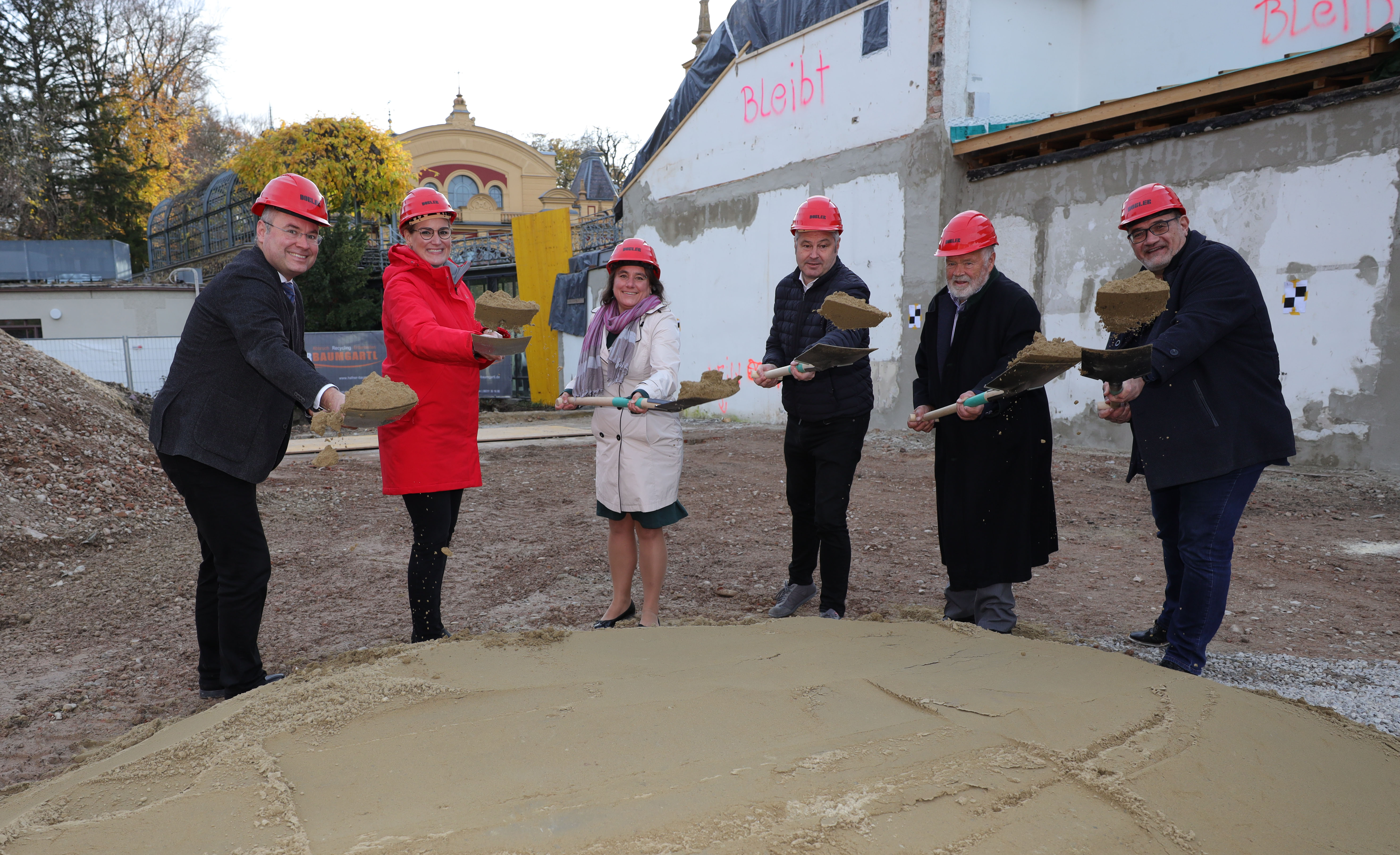 Sechs Personen mit Bauhelm auf dem Kopf und Spaten in den Händen schaufeln Sand von einem kleinen Hügel auf einer Baustelle.