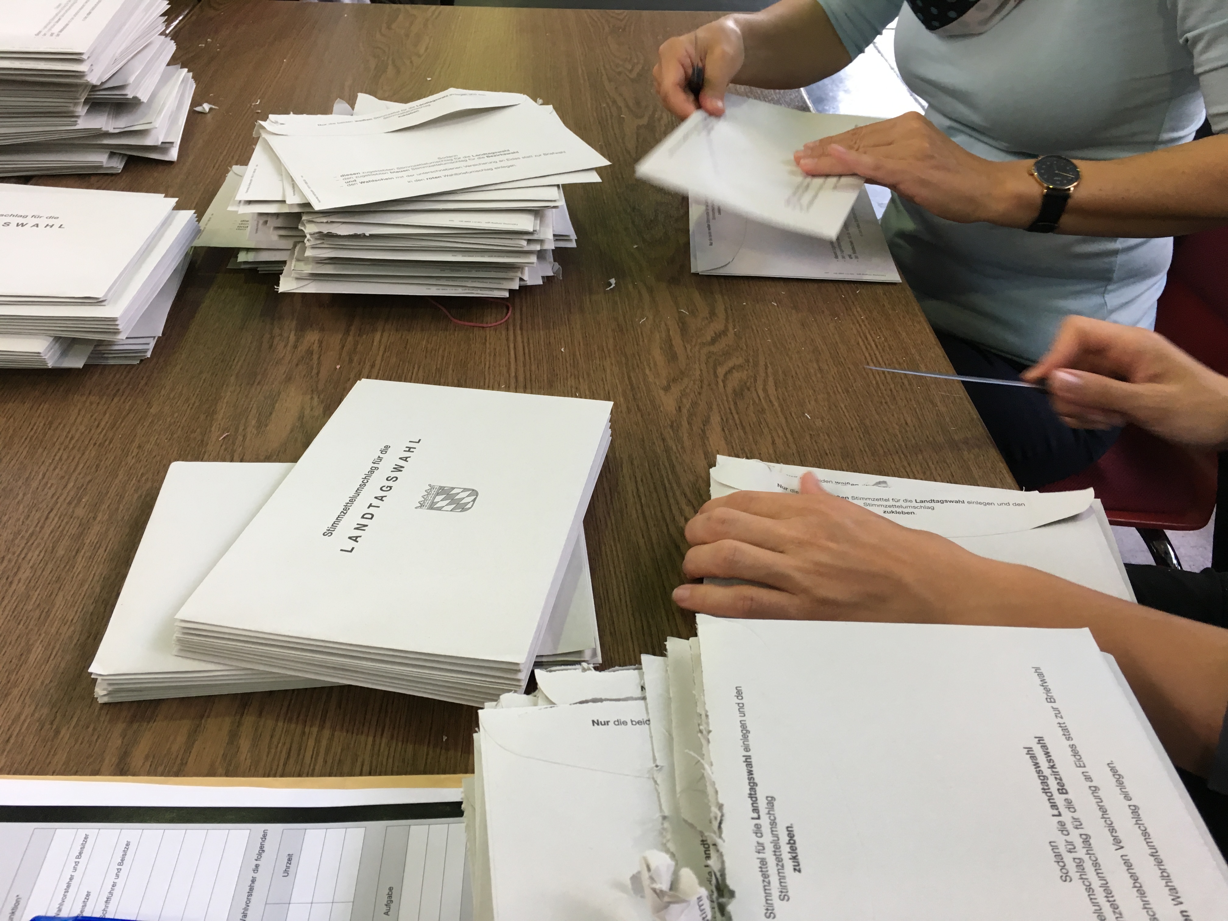 Auf einem Tisch liegen viele Wahlzettel. Rechts sind die Hände zweier Personen zu sehen. Sie öffnen je einen Briefumschlag. Auf den Umschlägen steht Landtagswahl 2018