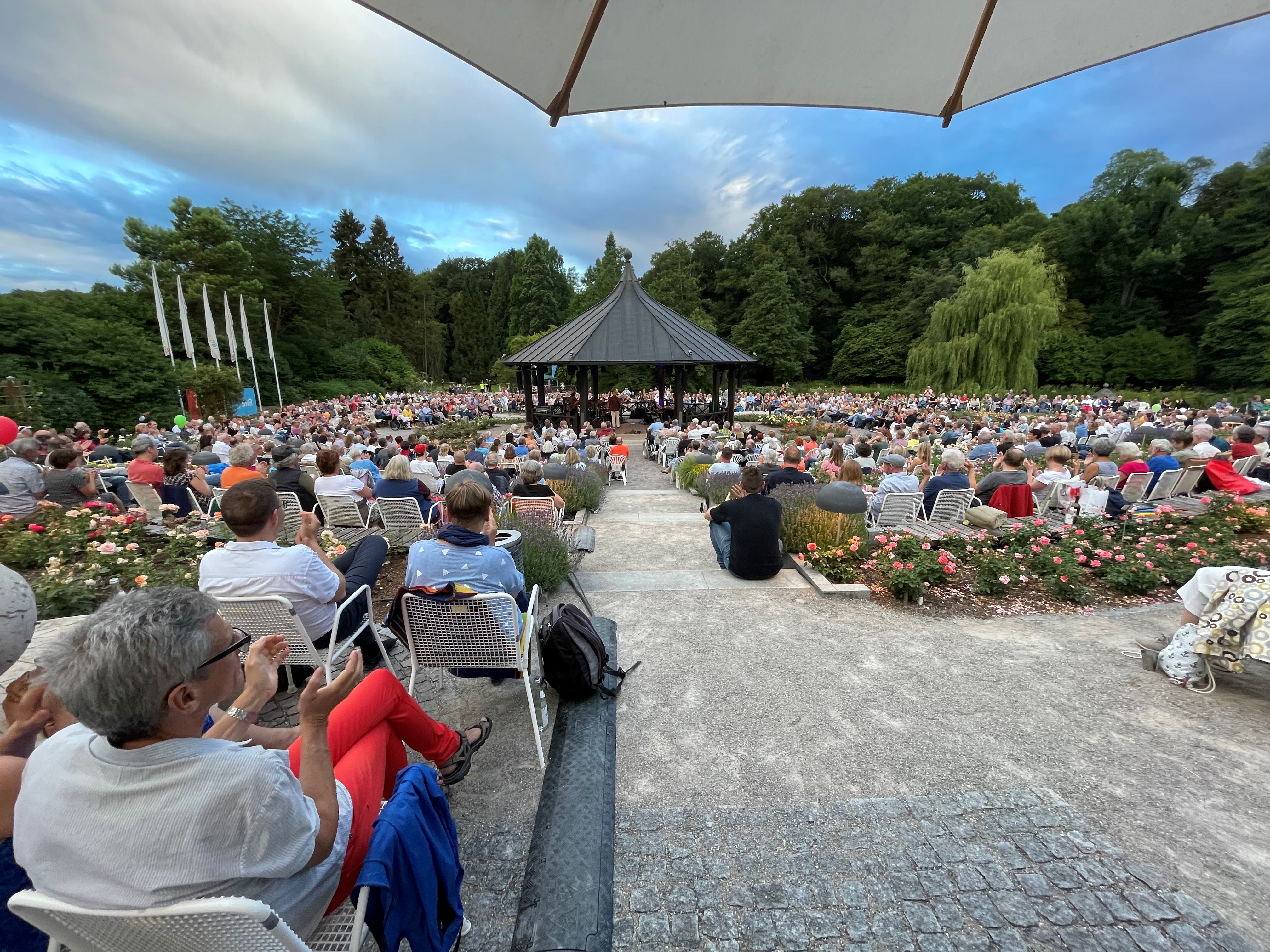 Totale vom Pavillon im Rosengarten, in dem ein Orchester spielt; umringt von Zuschauern in weißen Gartenstühlen