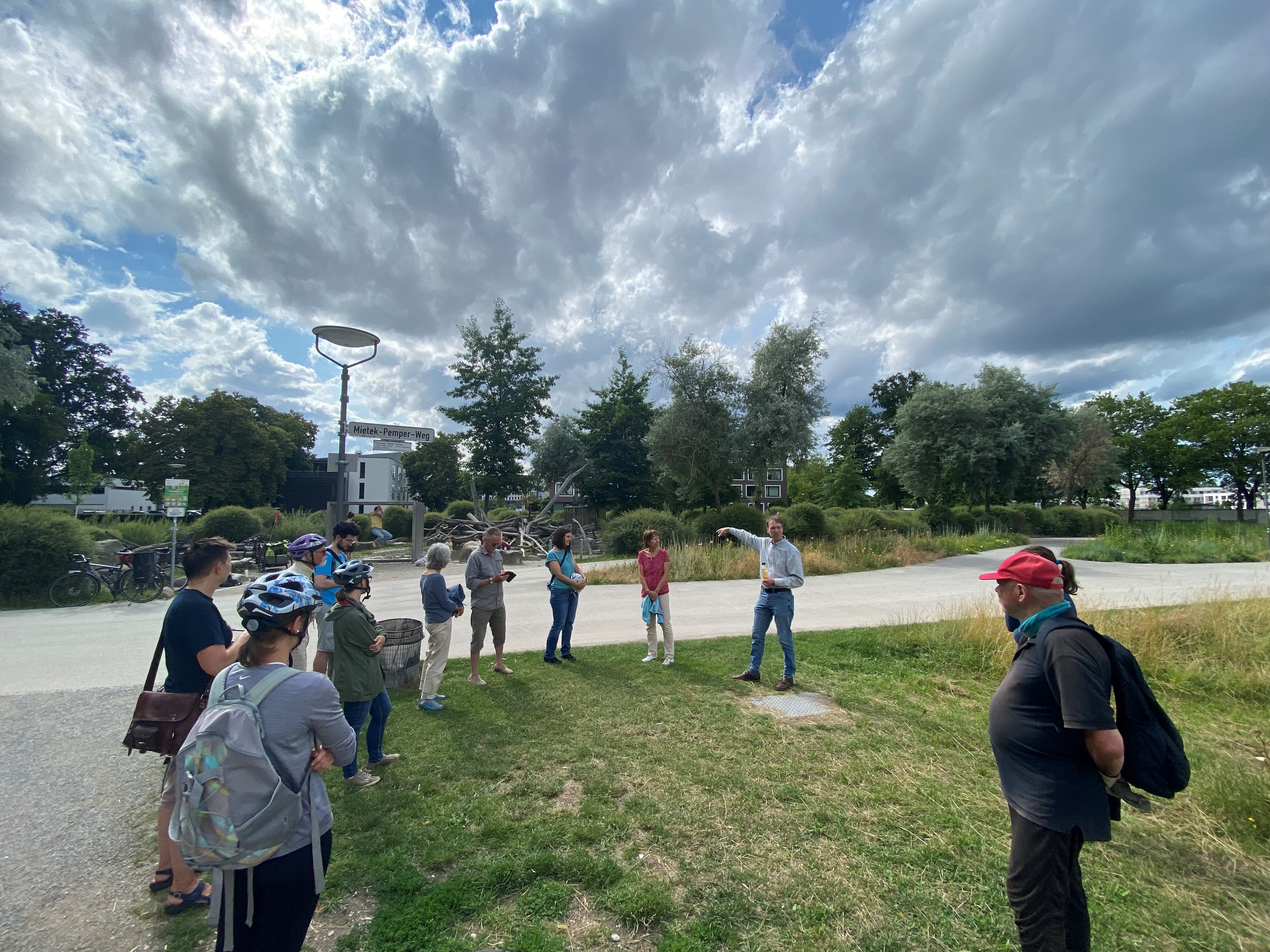 Fahrradtour „Klimawandel und Wasser“. Foto: Gregor Rudat/Stadt Augsburg Etwa zehn Erwachsene stehen auf einer Wiese. Im Hintergrund ist ein Spielplatz zu sehen. Dazwischen verläuft ein Fuß- und Radweg. Auf dem Straßenschild steht "Mietek-Pemper-Weg"