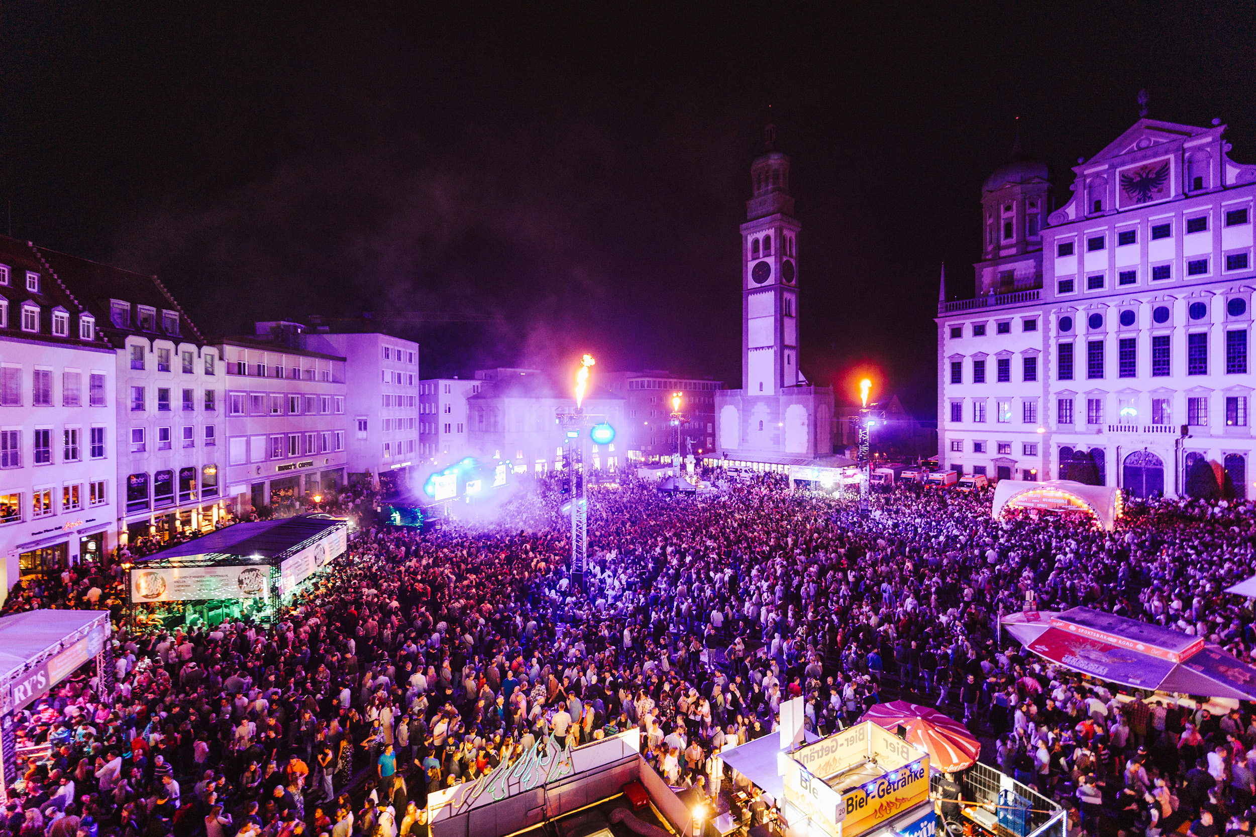 Der Augsburger Rathausplatz voller Menschen. Es ist dunkel und alles ist in pinkes Licht getaucht. Es gibt Buden und eine Bühne.