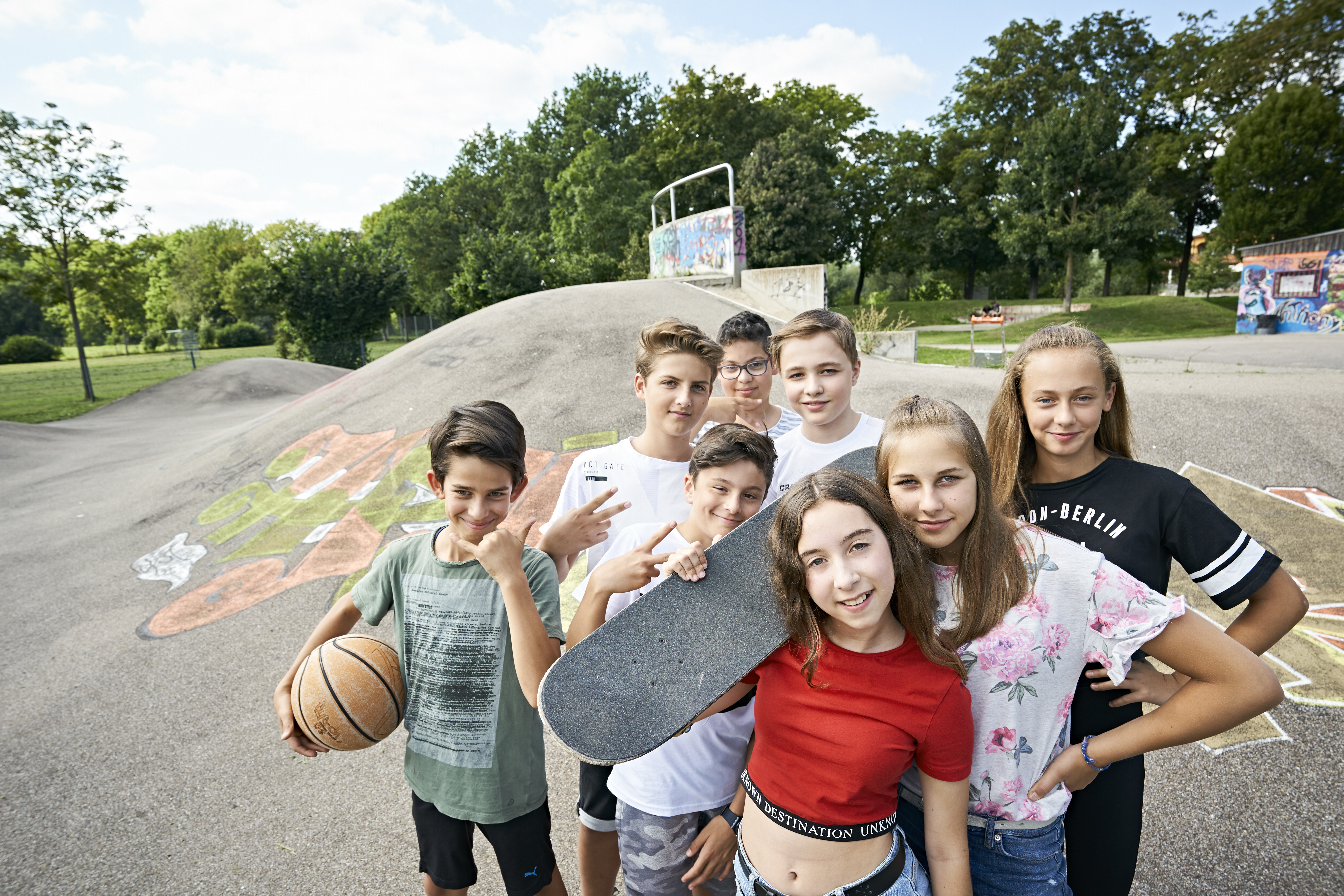 Acht Jugendliche stehen mit Skateboards und einem Basektball auf einer Skater-Anlage