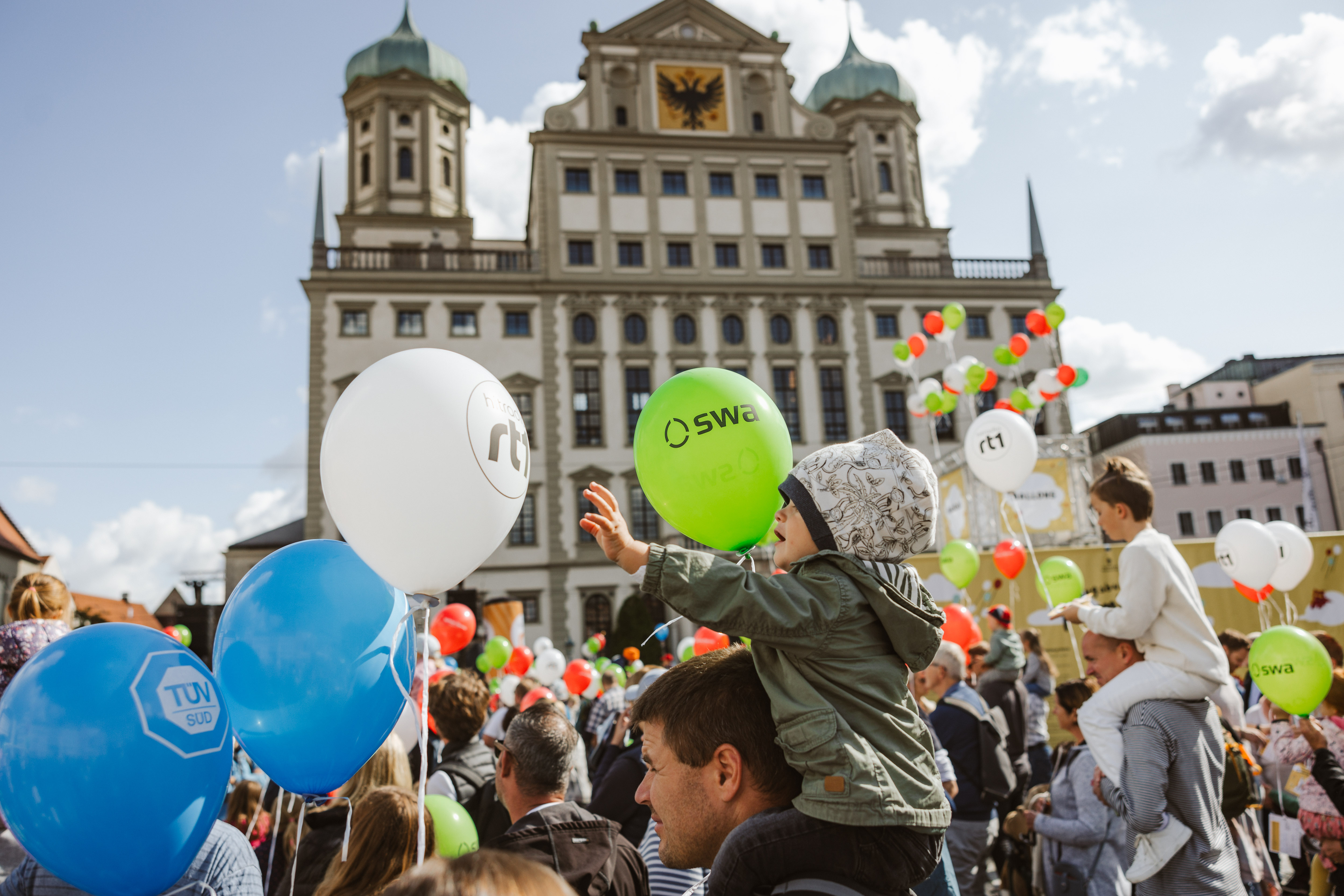 Augsburger Rathaus, im Voredergrund Eltern, die ihre Kinder auf den Schultern tragen; alle halten Luftballons
