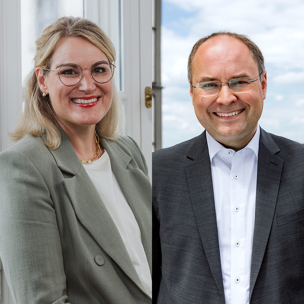 links eine Frau mit blondem, halblangen Haar mit Brille, rechts ein - ebenfalls bebrillter - Mann im Anzug