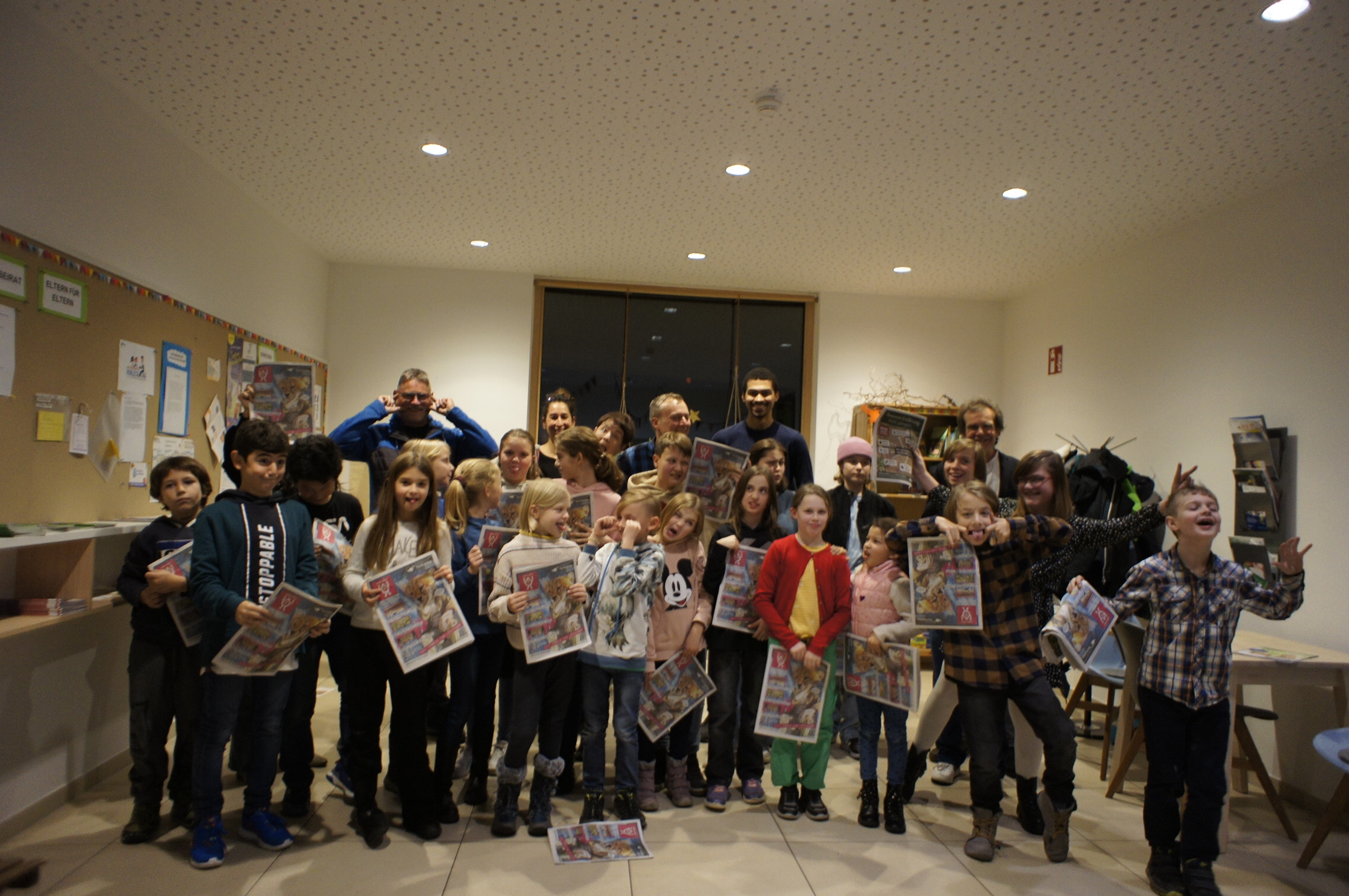 Etwa 20 Kinder und fünf Erwachsene halten bunte Zeitungen in der Hand. Viele Kinder machen Grimassen.