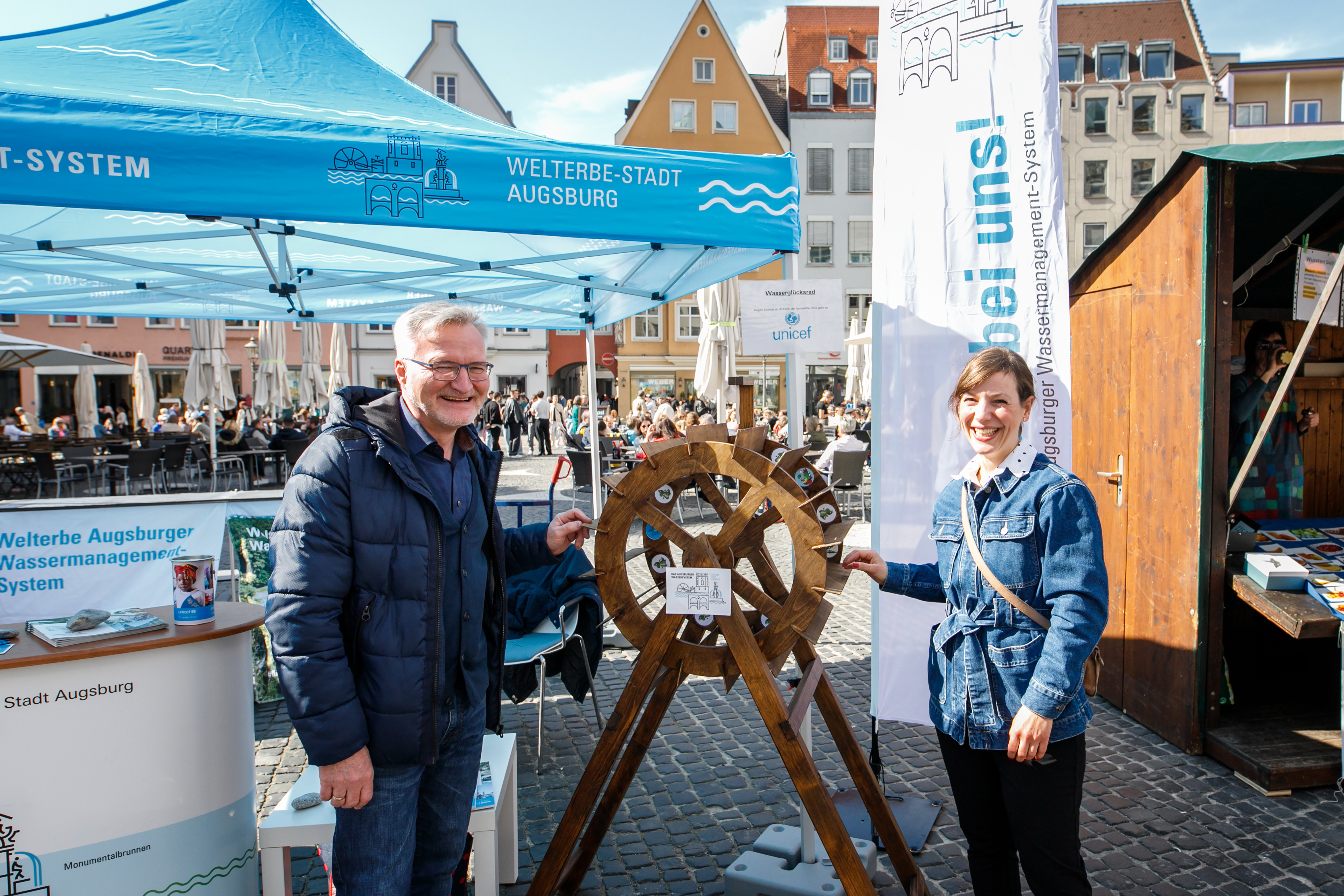 Ein Mann und eine Frau stehen auf dem Rathausplatz. Zwischen ihnen ein Modell eines Wasserrades aus Holz