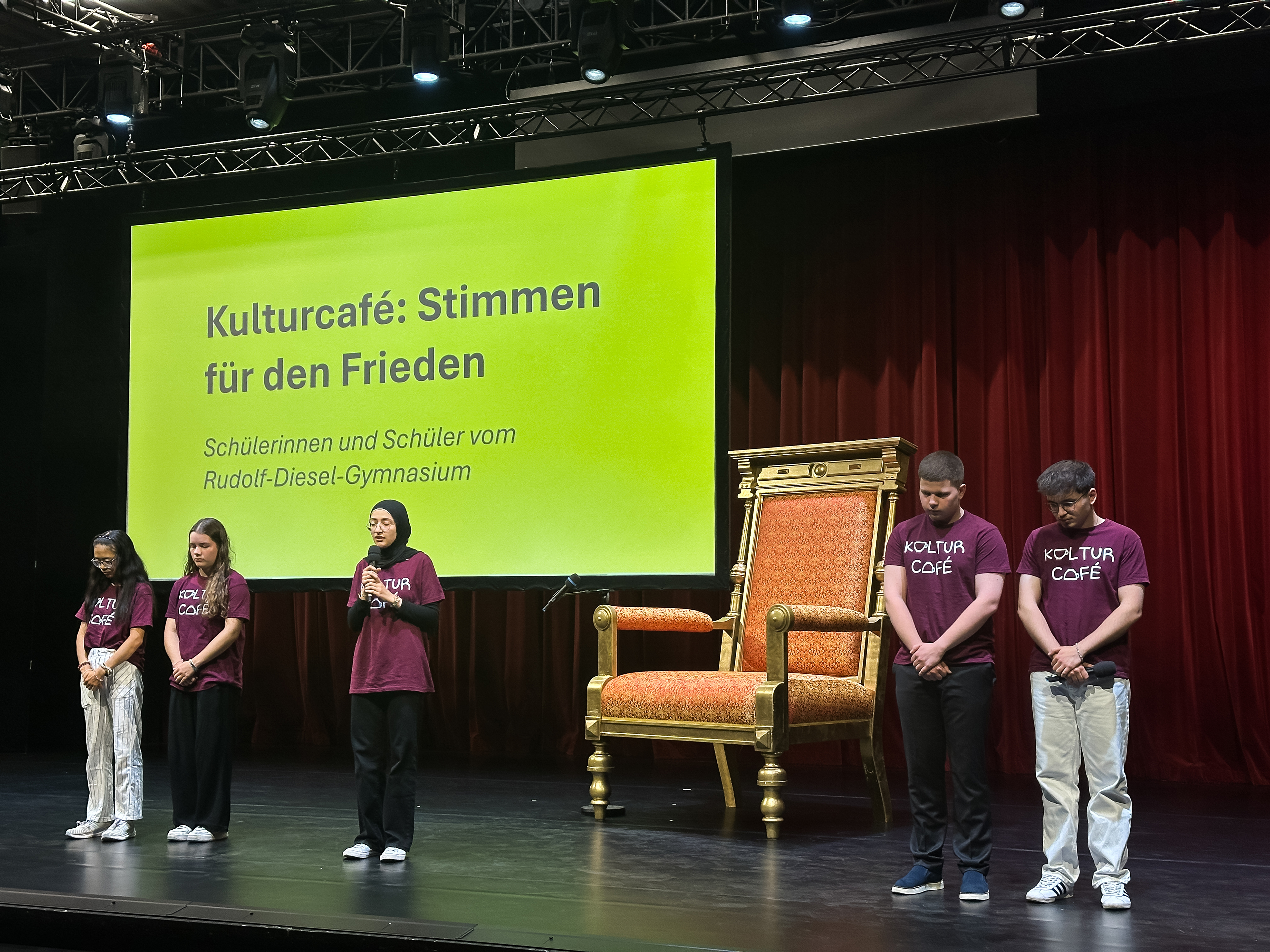 Fünf Schülerinnen und Schüler stehen auf einer Bühne. Im Hintergrund steht ein riesiger Stuhl. 
