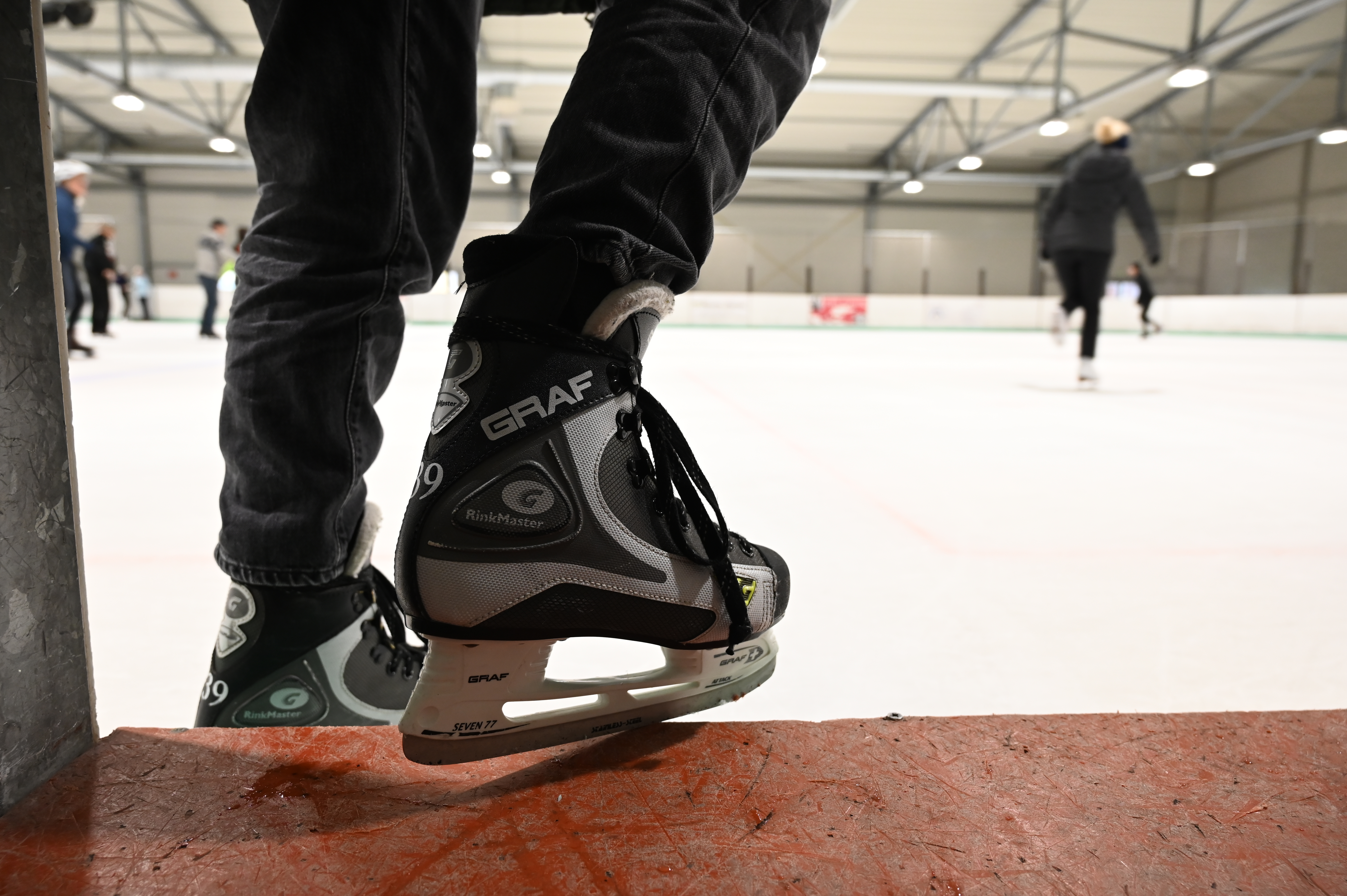 Im Vordergrund Füße in Schlittschuhen, die gerade auf die Eisfläche steigen. Im Hintergrund fahren mehrere Personen Schlittschuh in der Eishalle Haunstetten