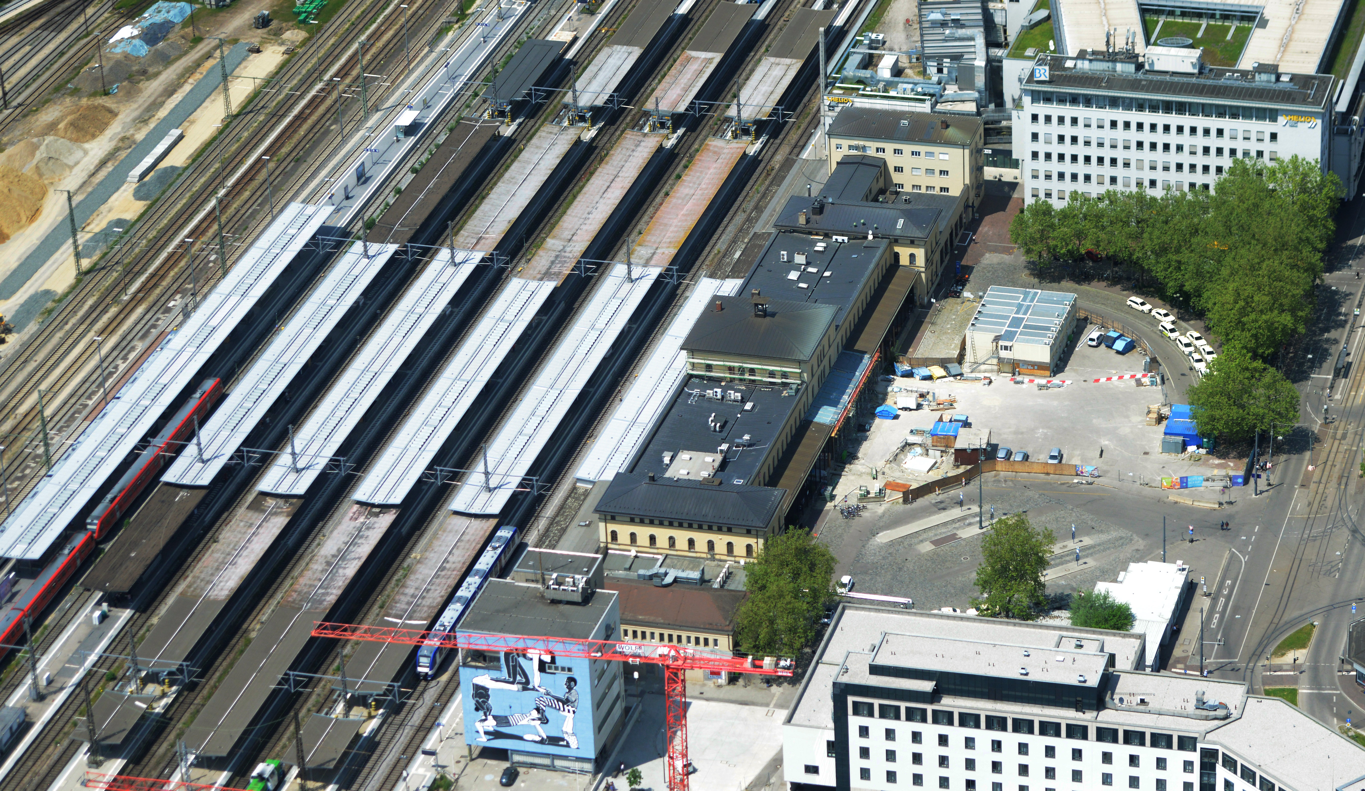 Auf dem Bild sieht man einen Bahnhof von oben. Rechts befindet sich der Bahnhofsvorplatz mit Bäumen. 