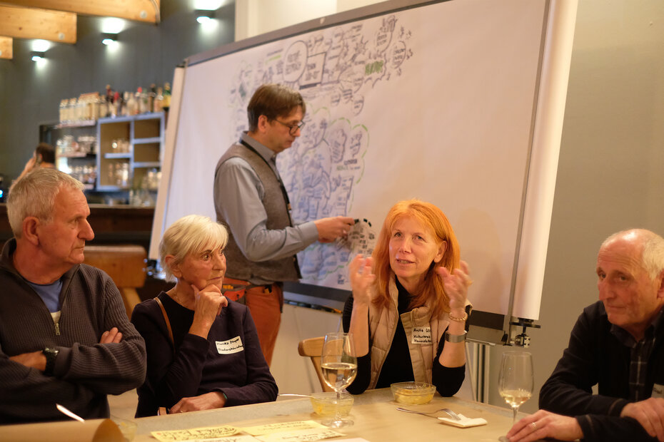 Im Vordergrund sitzen vier Personen an einem Disch und diskutieren. Im Hintergrund schreibt ein Mann auf eine weiße Tafel.