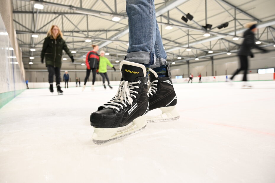 Ein Schlittschuh auf dem Eis. Im Hintergrund Menschen, die Eislaufen