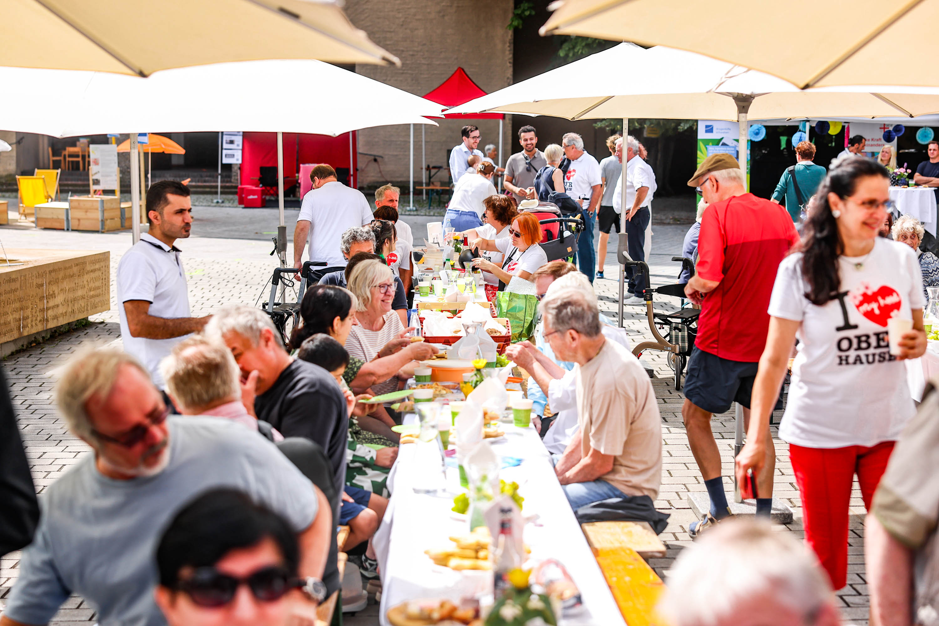 viele Leute sitzen unter Sonnenschirmen an Biertischgarnituren und lachen in die Kamera