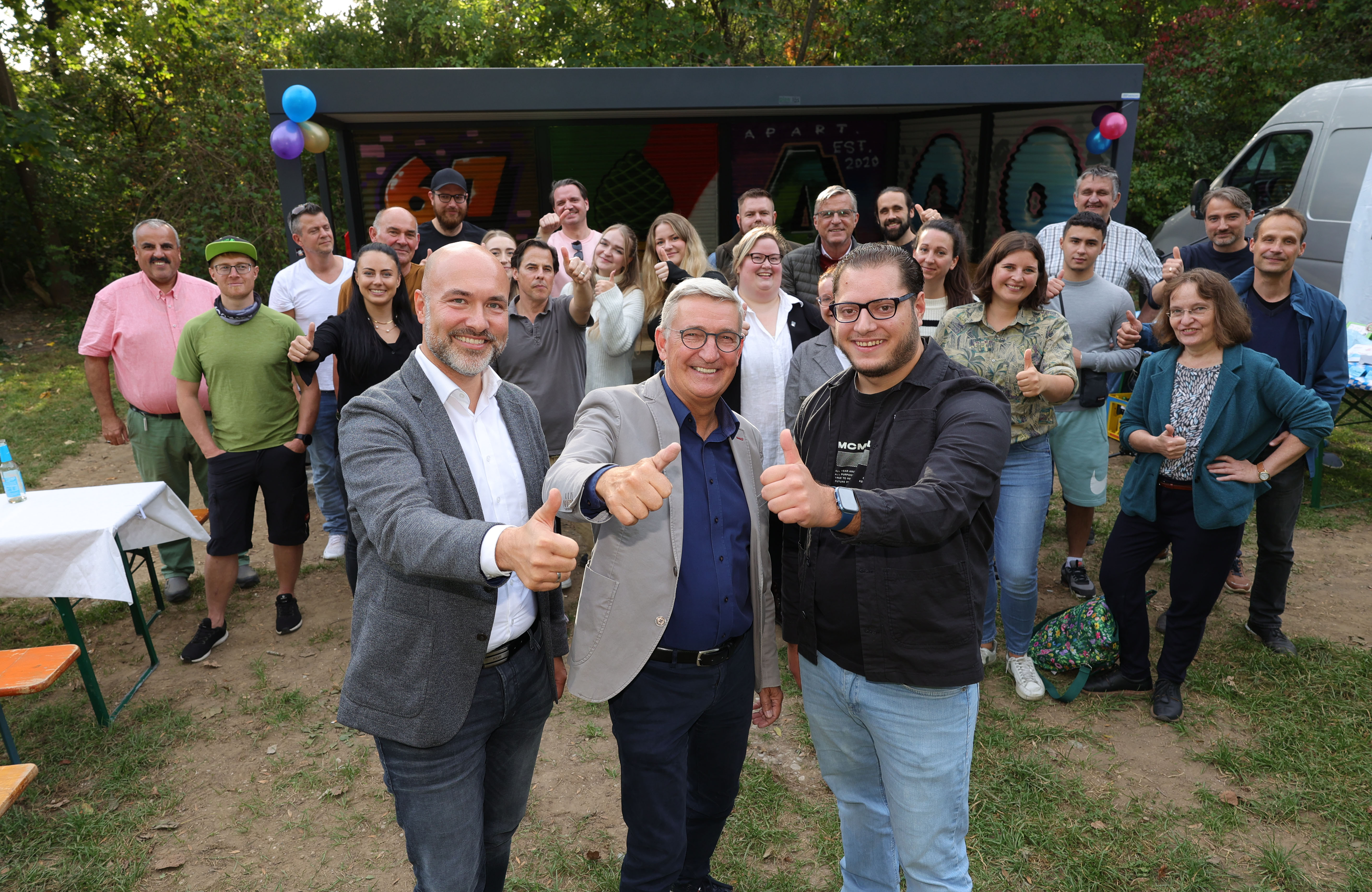 Viele Personen stehen vor einem Unterstand, der mit Luftballons geschmückt ist. Im Vordergrund befinden sich drei Männer, die den Daumen nach oben zeigen. Im Hintergrund stehen viele Menschen, die ebenfalls den Daumen nach oben zeigen bzw. in die Kamera lächeln.