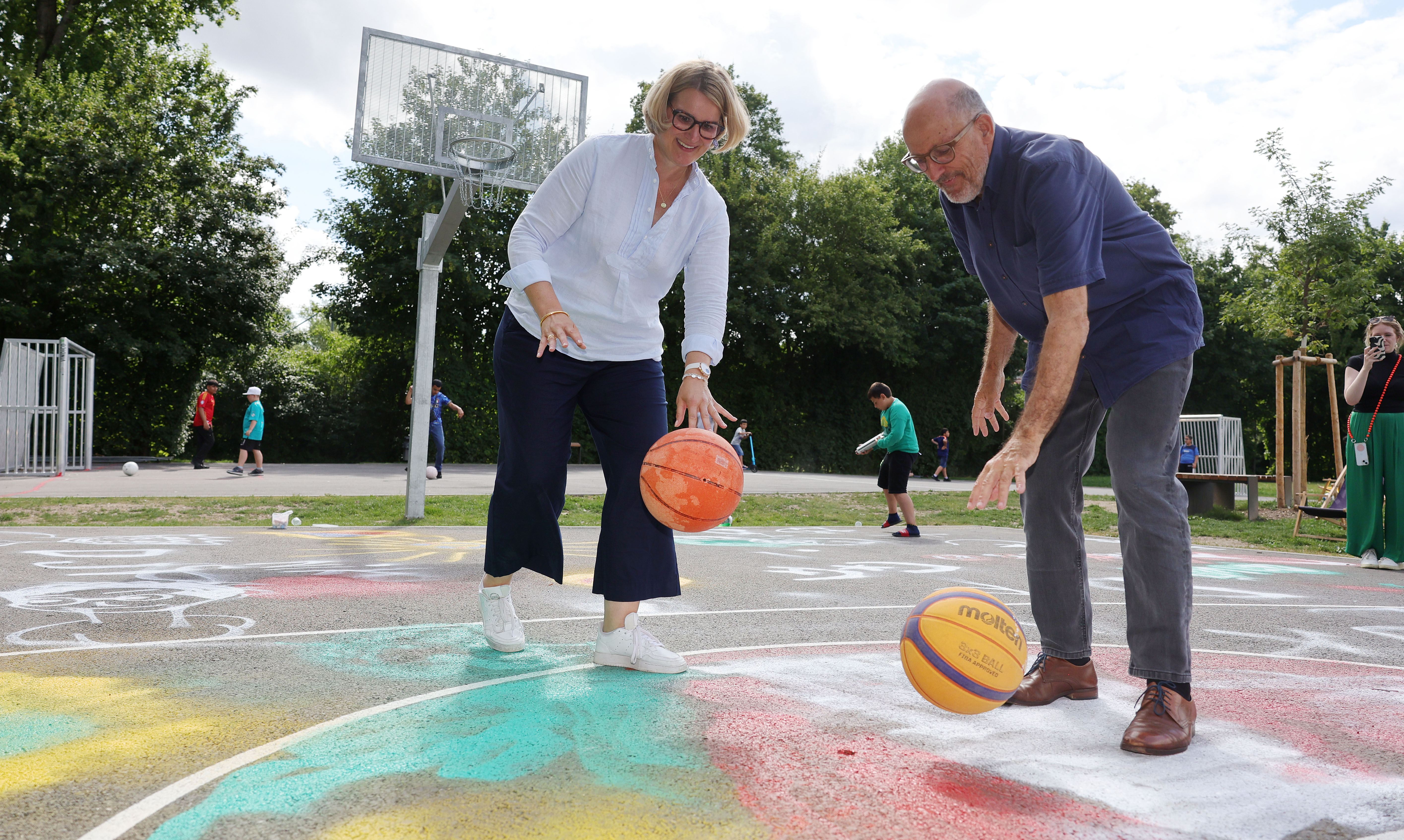 Eine Frau und ein Mann dribbeln Basketbälle auf einem SPortplatz. Der Boden ist geteert und bemalt. Im Hintergrund sieht man einen Basketballkorb