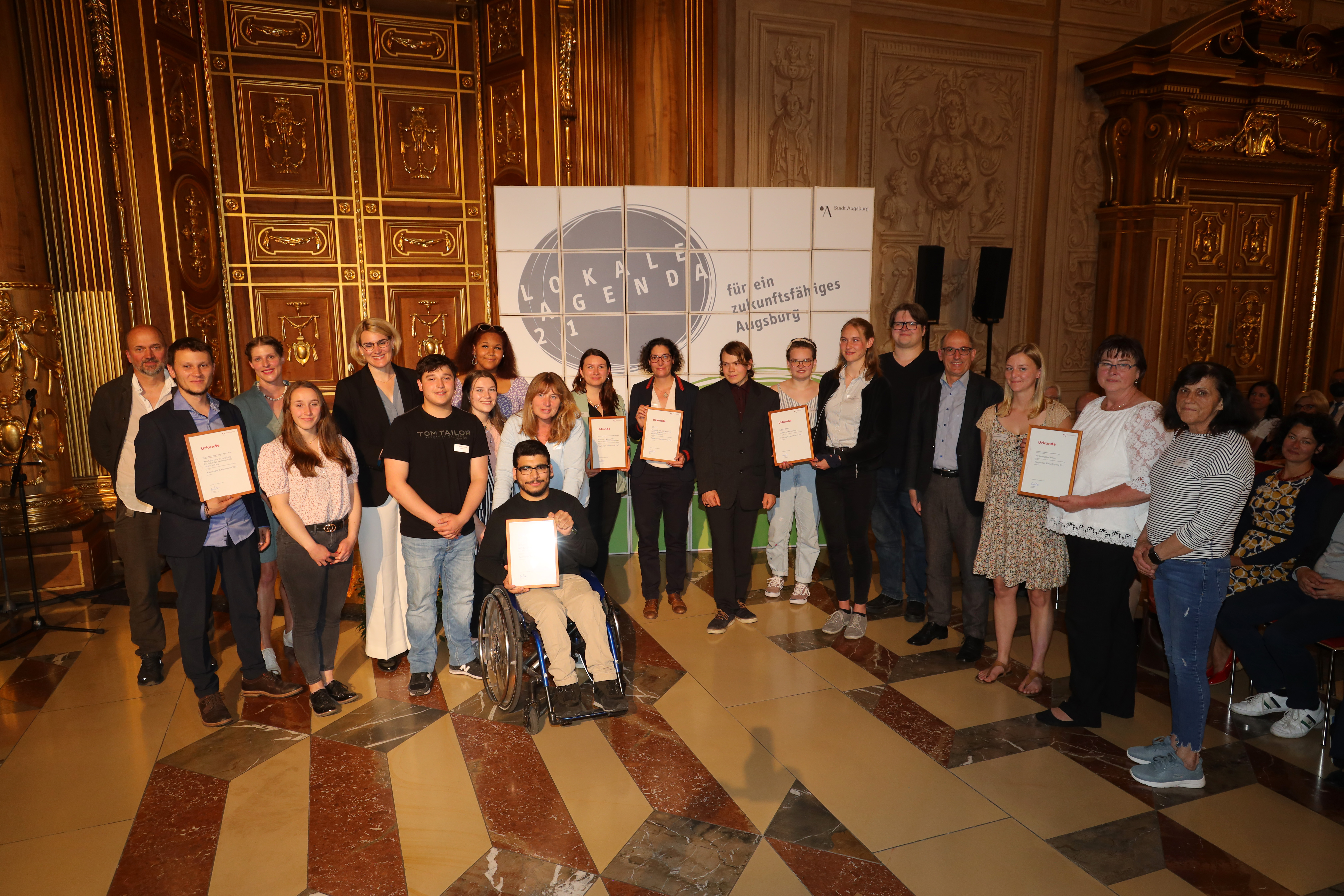 Rund 25 Personen stehen im Goldenen Saal vor einer Wand mit der Aufschrift "Lokale Agenda 21". Einige Personen haben Urkunden in der Hand.