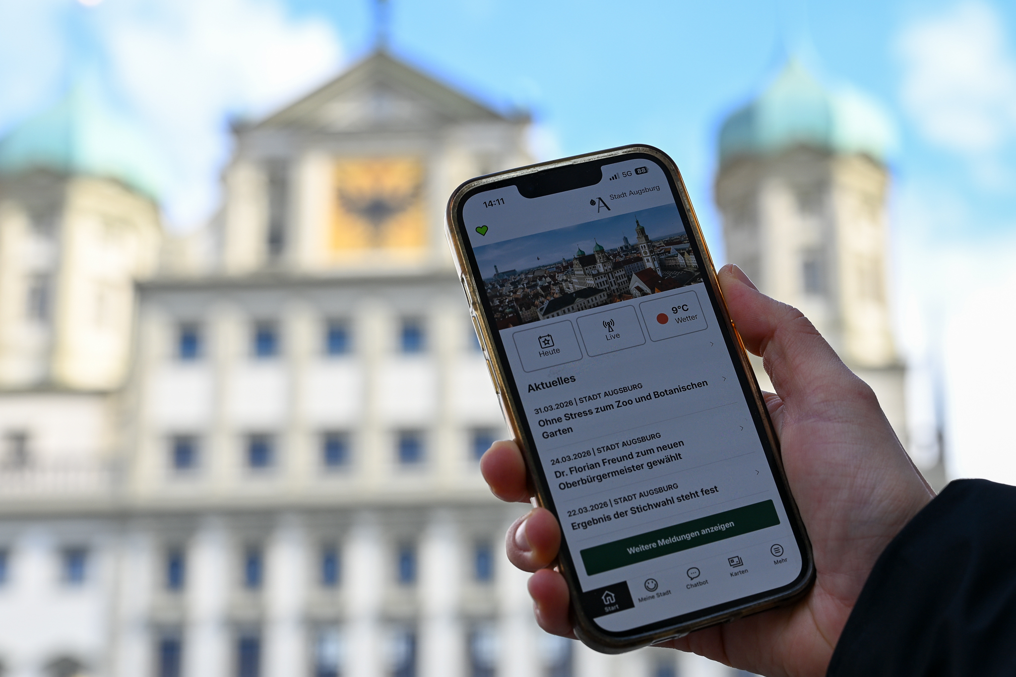 Zu sehen ist eine Hand, die ein Smartphone hält. Darauf ist die Augsburg App zu sehen. Im Hintergrund  das Augsburger Rathaus
