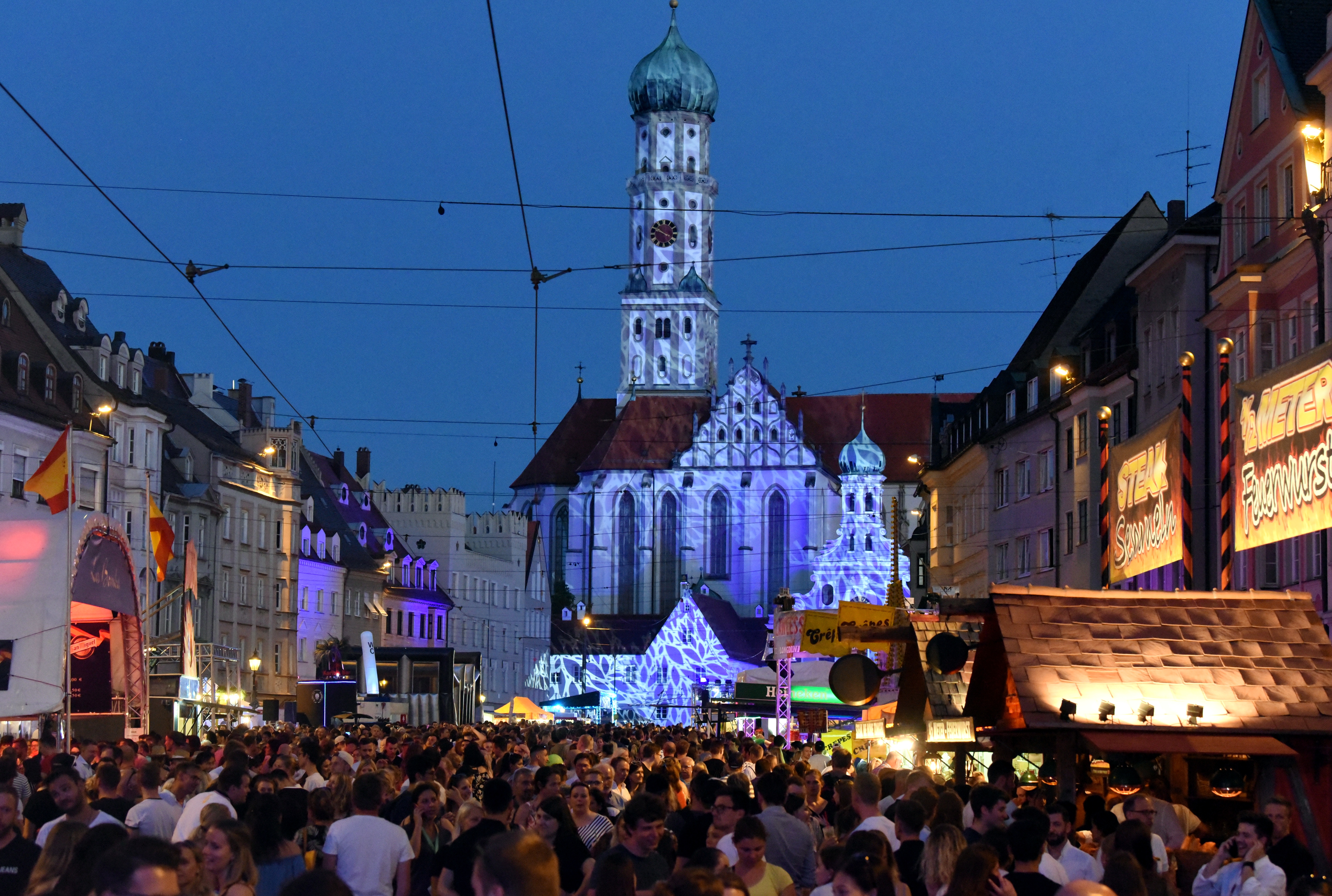 In der Maximilianstraße feiern viele Menschen. Es ist Abend. Die Kirche St. Ulrich ist blau beleuchtet.