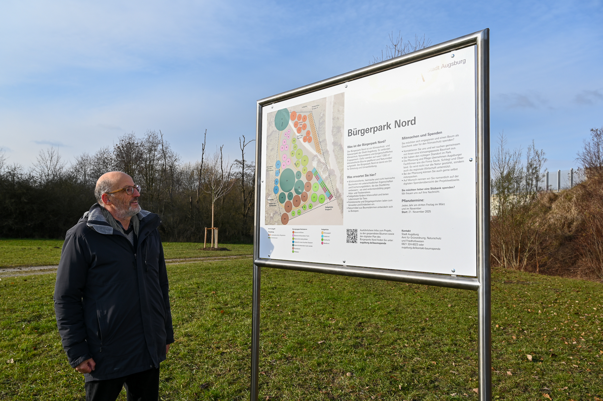 Im Vordergrund steht Umweltreferent Reiner Erben neben einer Informationstafel zum Bürgerpark Nord, im Hintergrund ein frisch gepflanzter, unbelaubter Baum