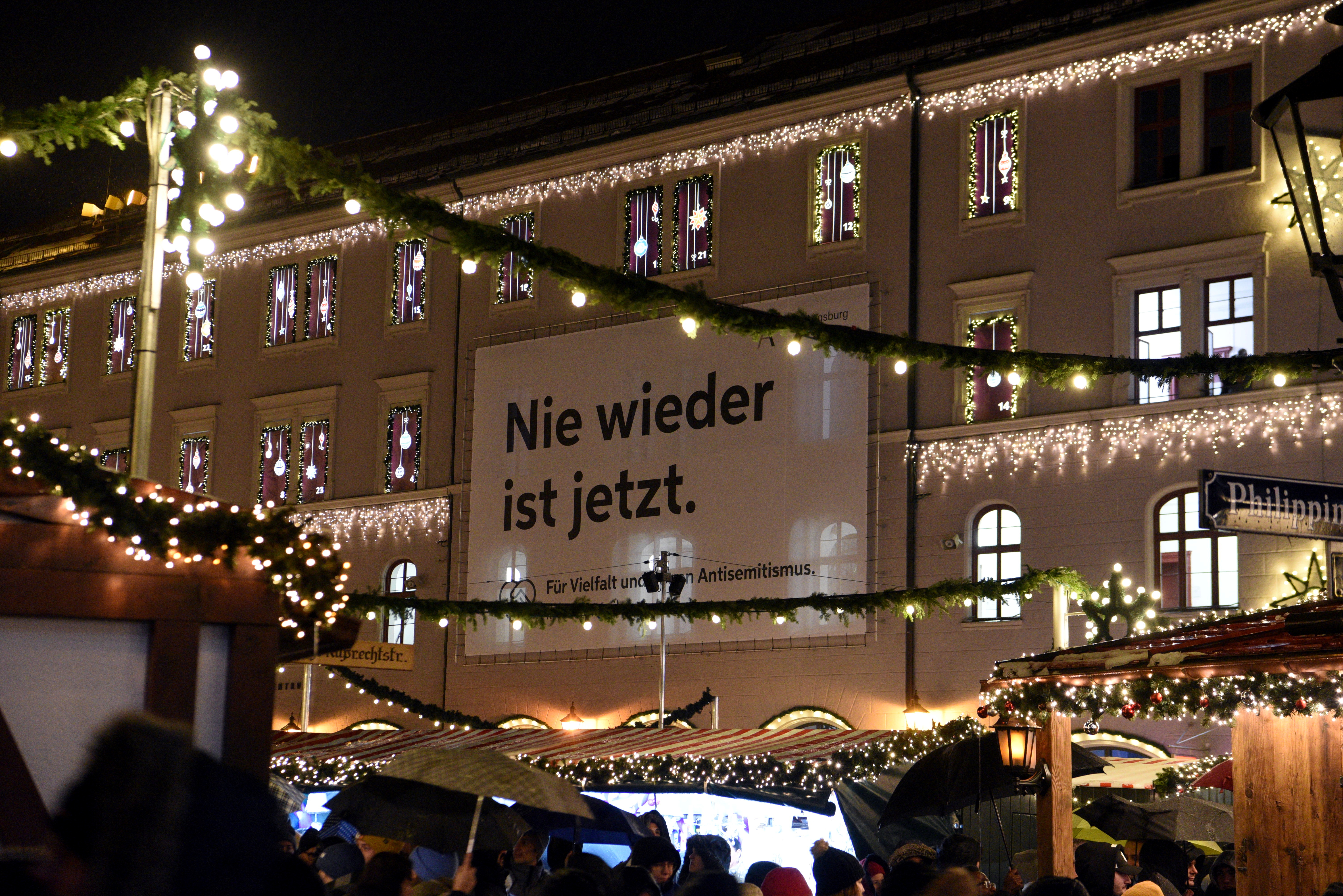 Der Banner mit dem Text "Nie wieder ist jetzt" am Verwaltungsgebäude 1 auf dem Rathausplatz zwischen dem Adventskalender in den Fenstern des Gebäudes.