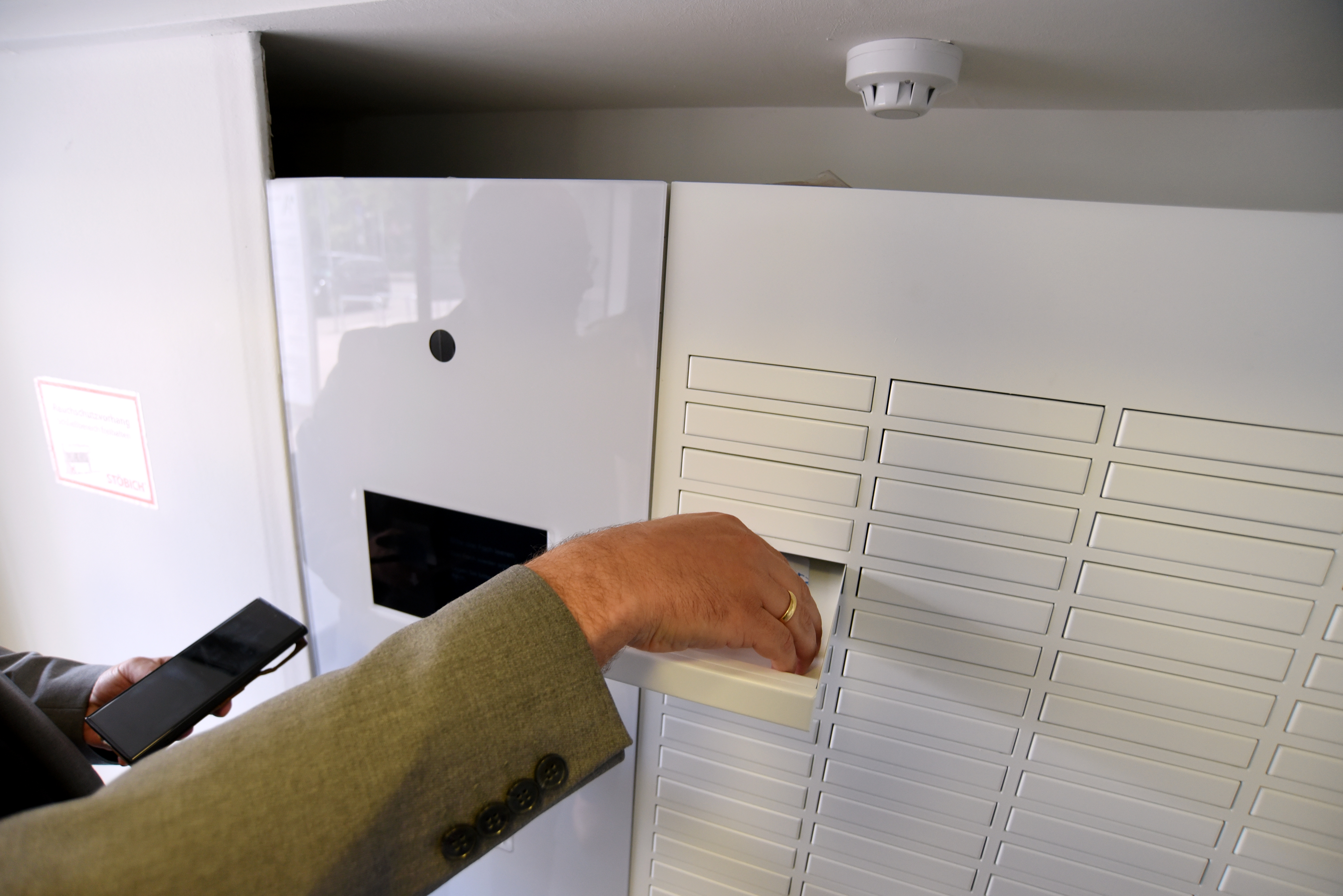 Ein Automat mit vielen kleinen Schubladen. Eine Hand hält ein Handy, eine andere Hand öffnet einer Schublade