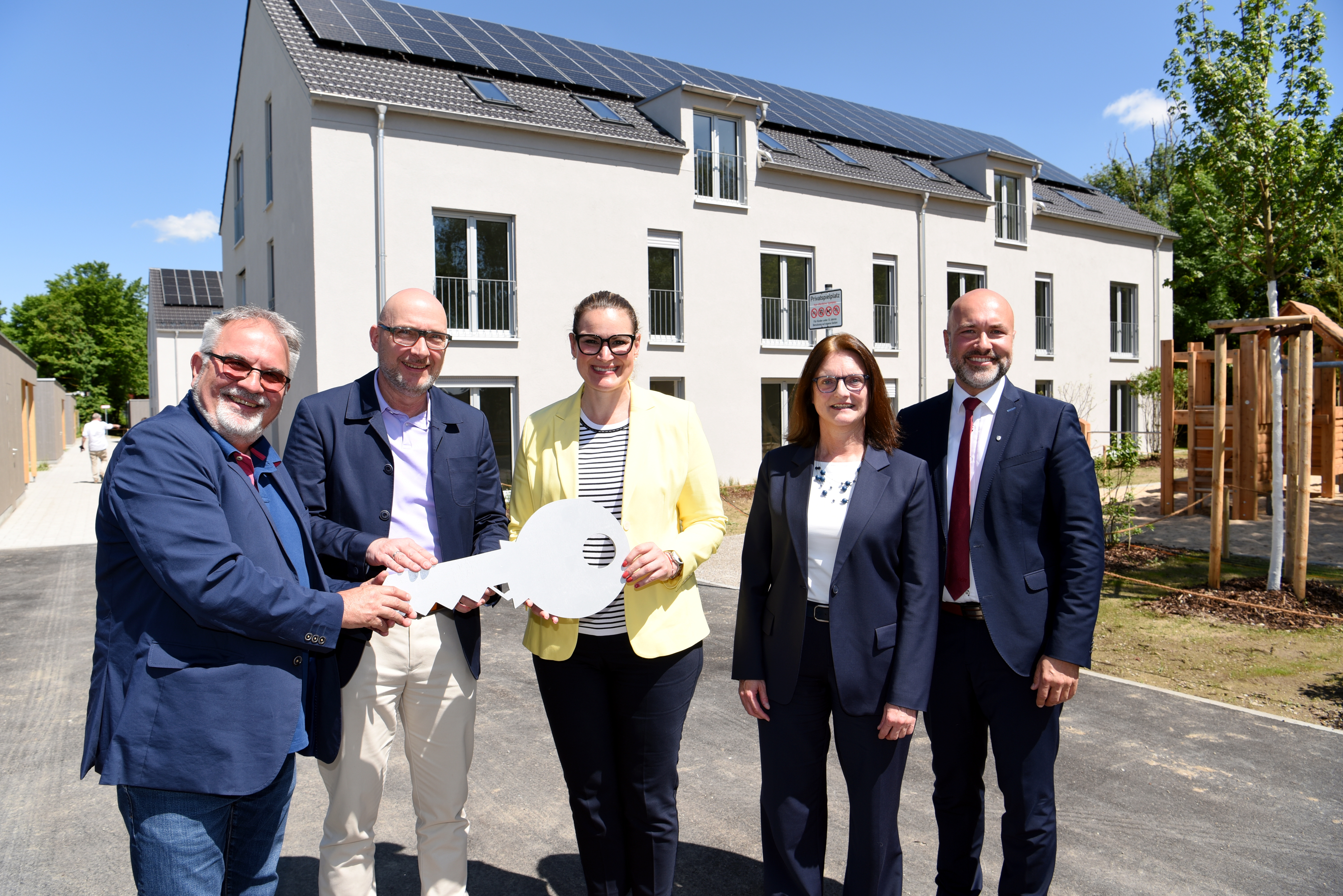 Fünf Menschen stehen vor einem Wohnhaus. Sie halten einen übergroßen Schlüssel in den Händen