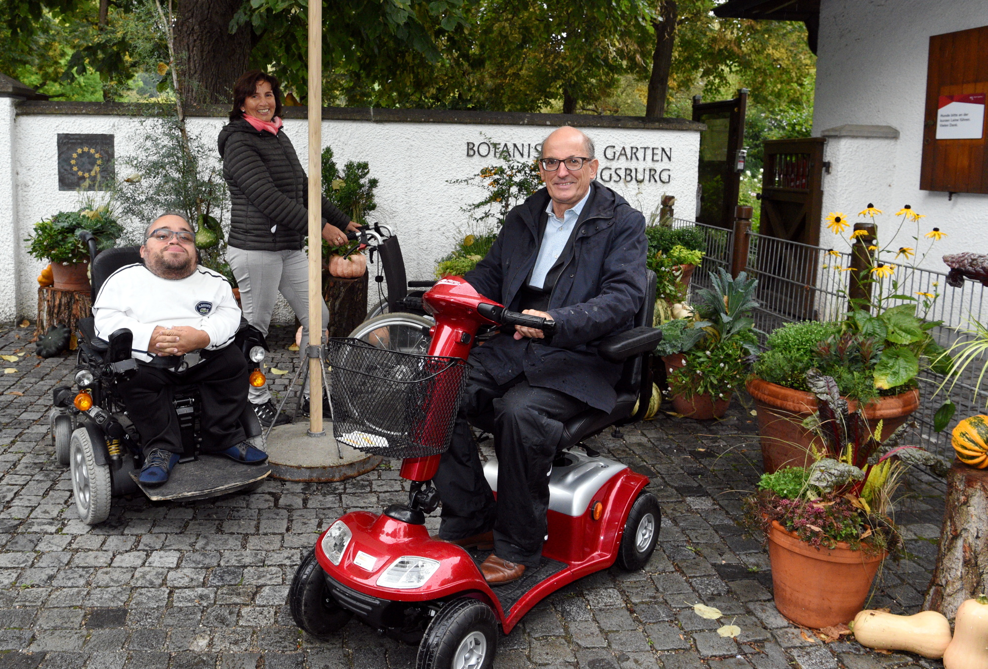 Drei Menschen mit verschiedenen Rollstühlen vor dem Tor des Botanischen Gartens. Von links Stadtrat Benedikt Lika im elektrischen Rollstuhl. In der Mitte eine Mitarbeiterin des Botanischen Gartens mit einem normalen Rollstuhl. Rechts Umweltreferent Reiner Erben mit dem neuen Elektro-Mobil.