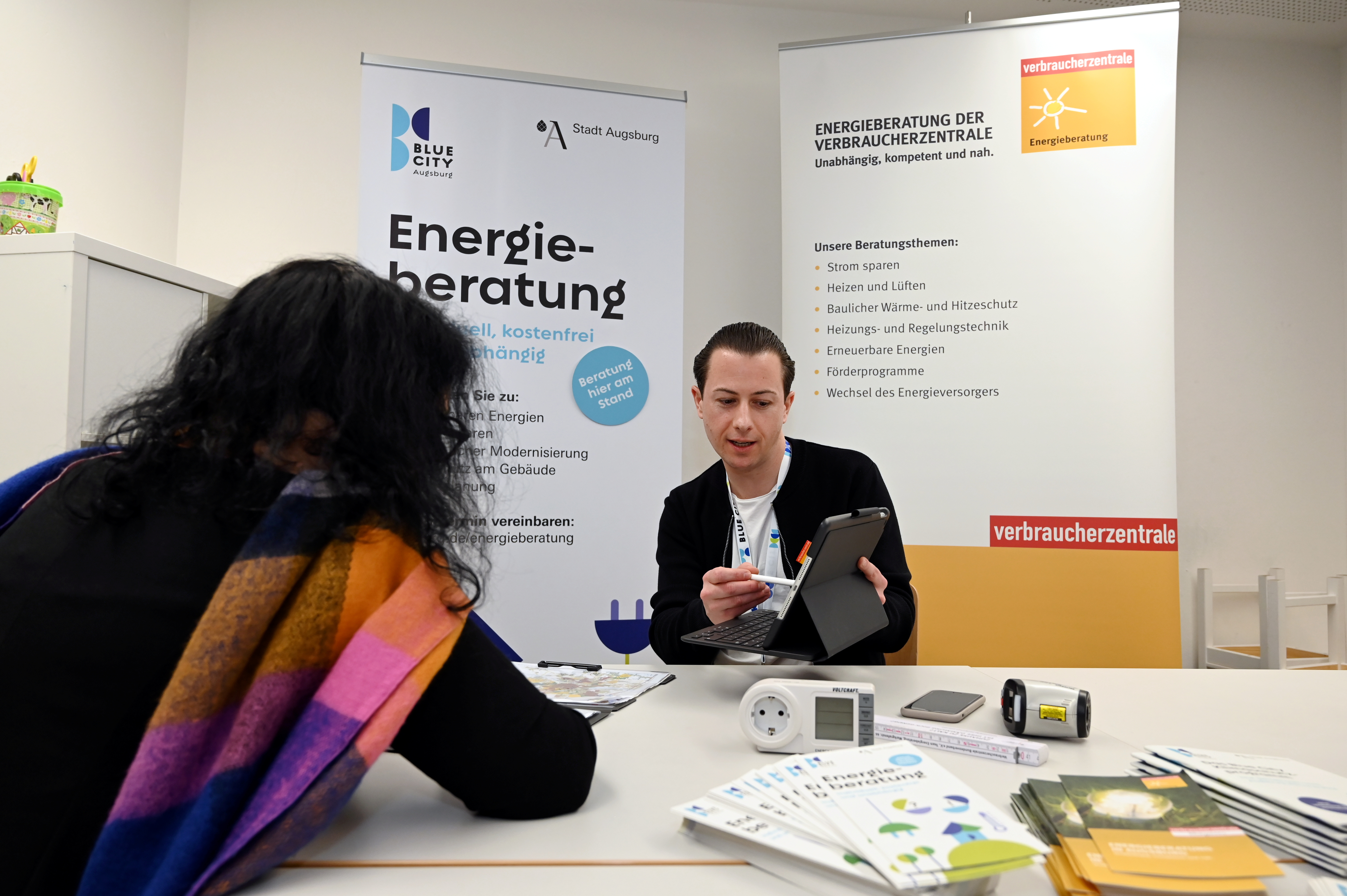 Ein Mann berät eine Frau. Im Hintergrund steht auf einem Plakat das Wort Energieberatung