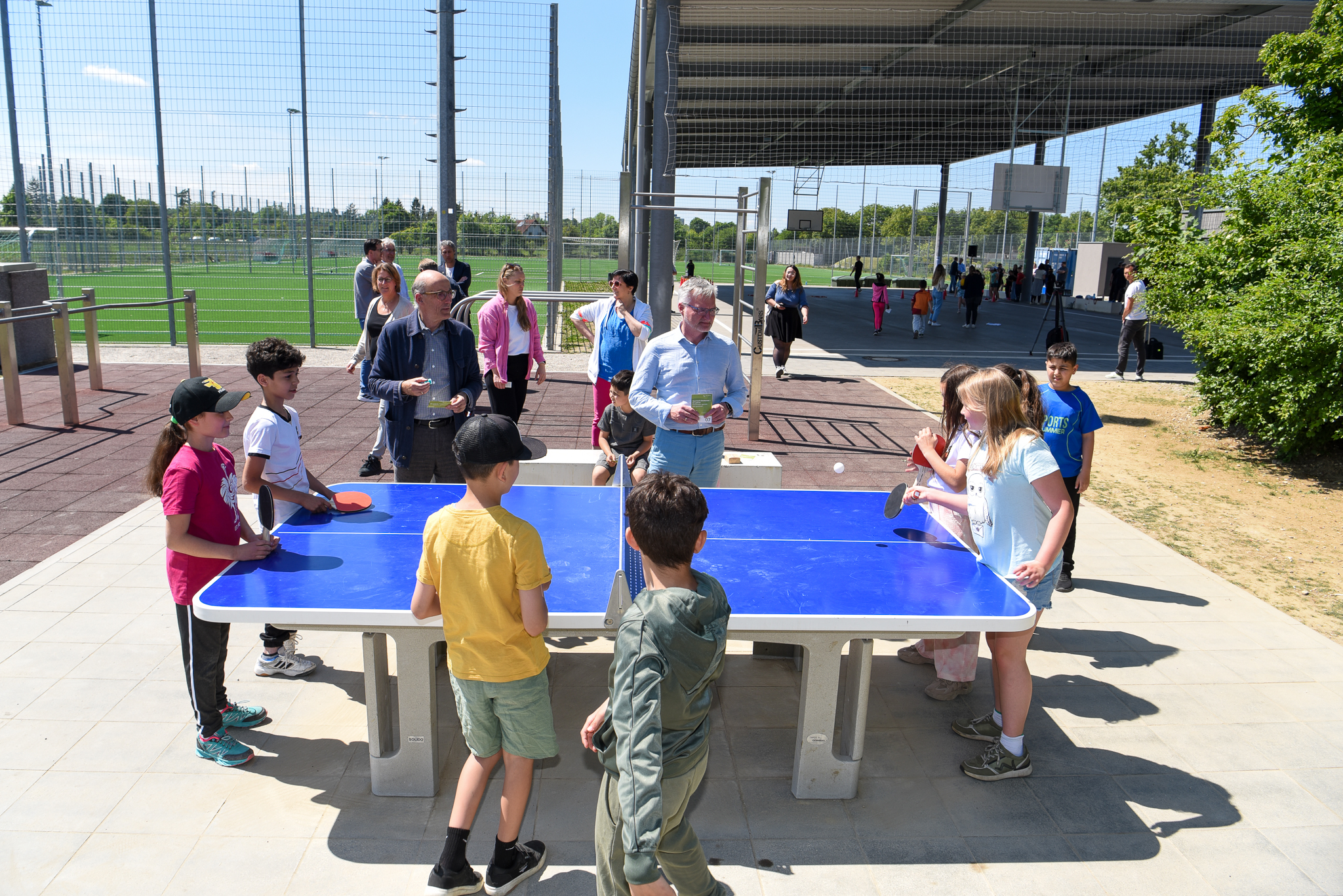Im Vodergrund viele Kinder an einer Tischtennisplatte. Im Hintergrund eine überdachte Basketball-Fläche und Fußballplätze