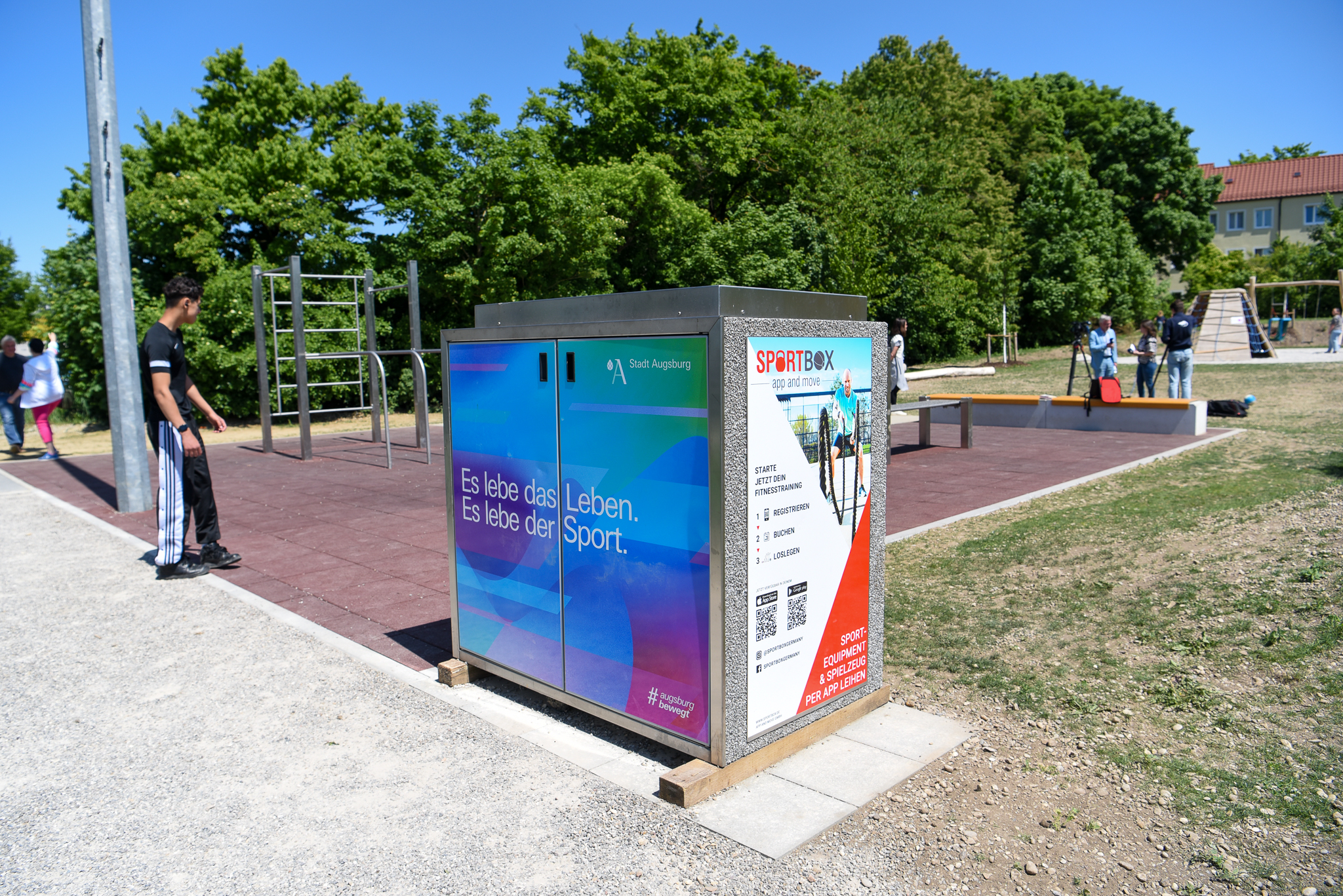 mannshohe Box mit rechteckigem Querschnitt, dahinter ein Spielfeld und Sportgeräte