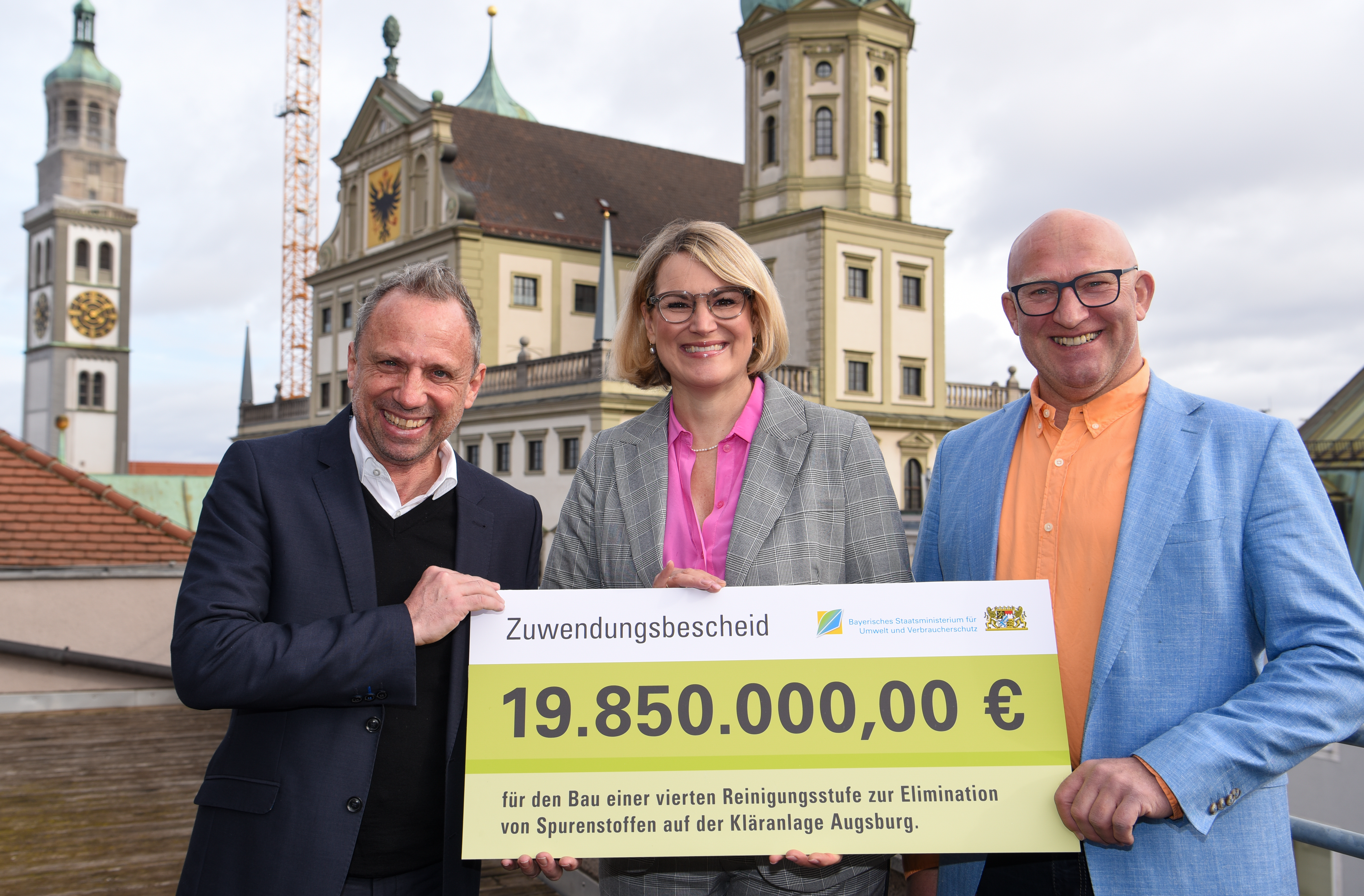 Zwei Männer und eine Frau halten ein Schild. Auf diesem steht das Wort "Zuwendungsbescheid" und der Geldbetrag 19,85 Millionen Euro