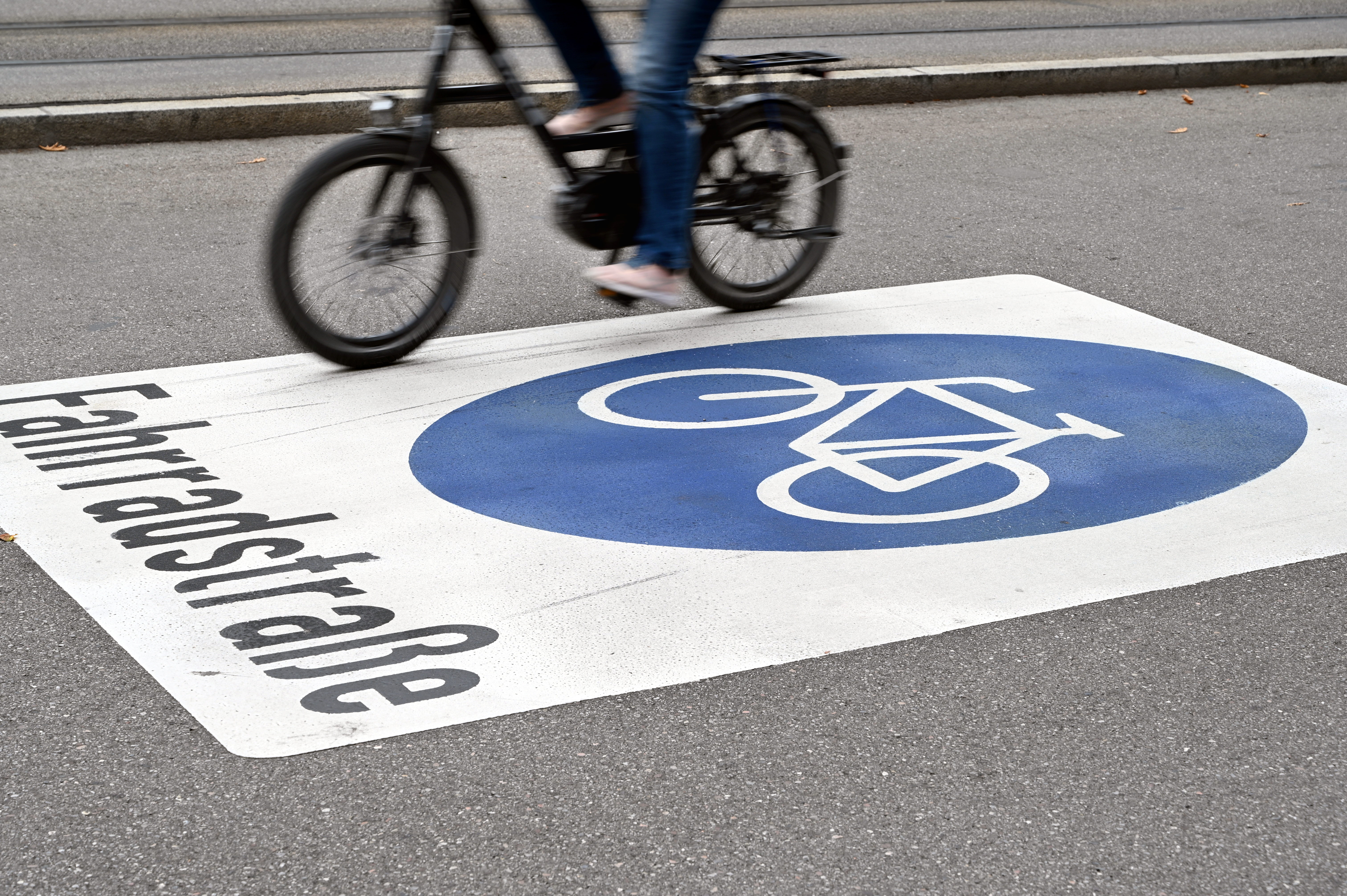 Auf einer Straße ist das Wort "Fahrradstraße" zu lesen, darüber ist ein blaues Radweg-Symbol gemalt. Ein Radfahrer fährt auf der Straße