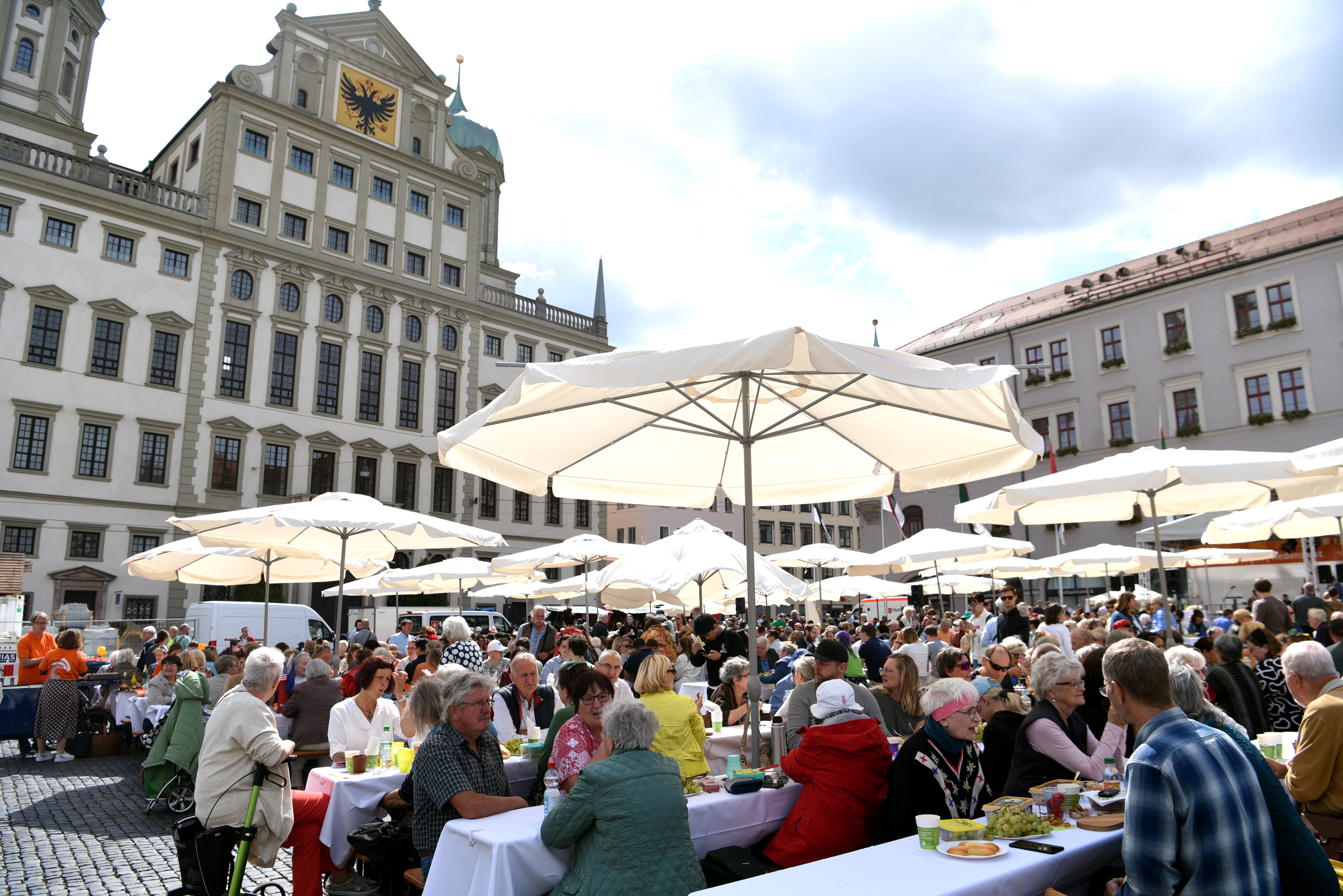 vor dem augsburger rathaus sitzen viele menschen unter sonnenschirmen an tischen zusammen