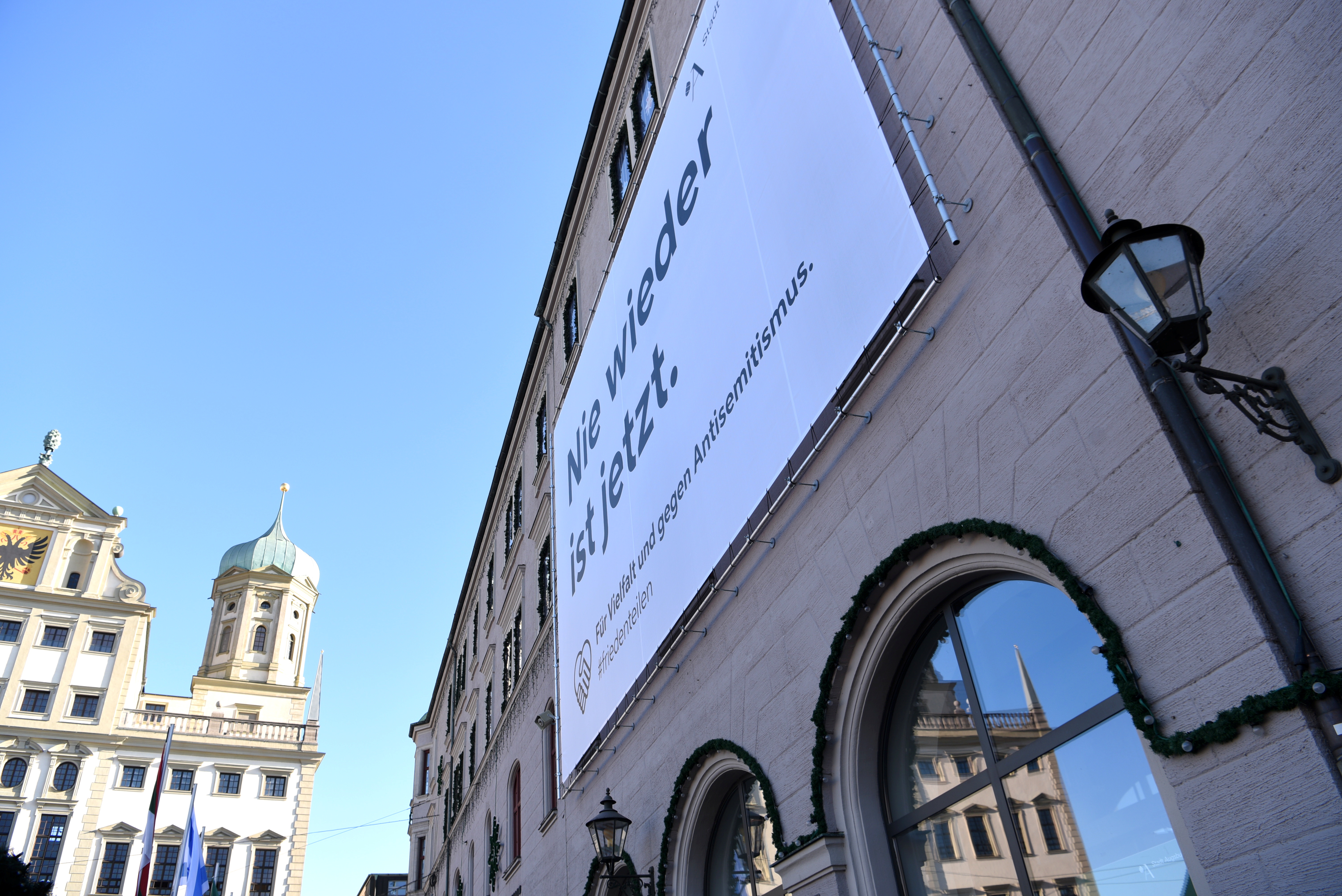 Ein großes Plakat mit der Aufschrift „Nie wieder ist jetzt. Für Vielfalt und gegen Antisemitismus“ hängt an der Fassade des Verwaltungsgebäudes. Am linken Bildrand ist das Rathaus zu sehen.