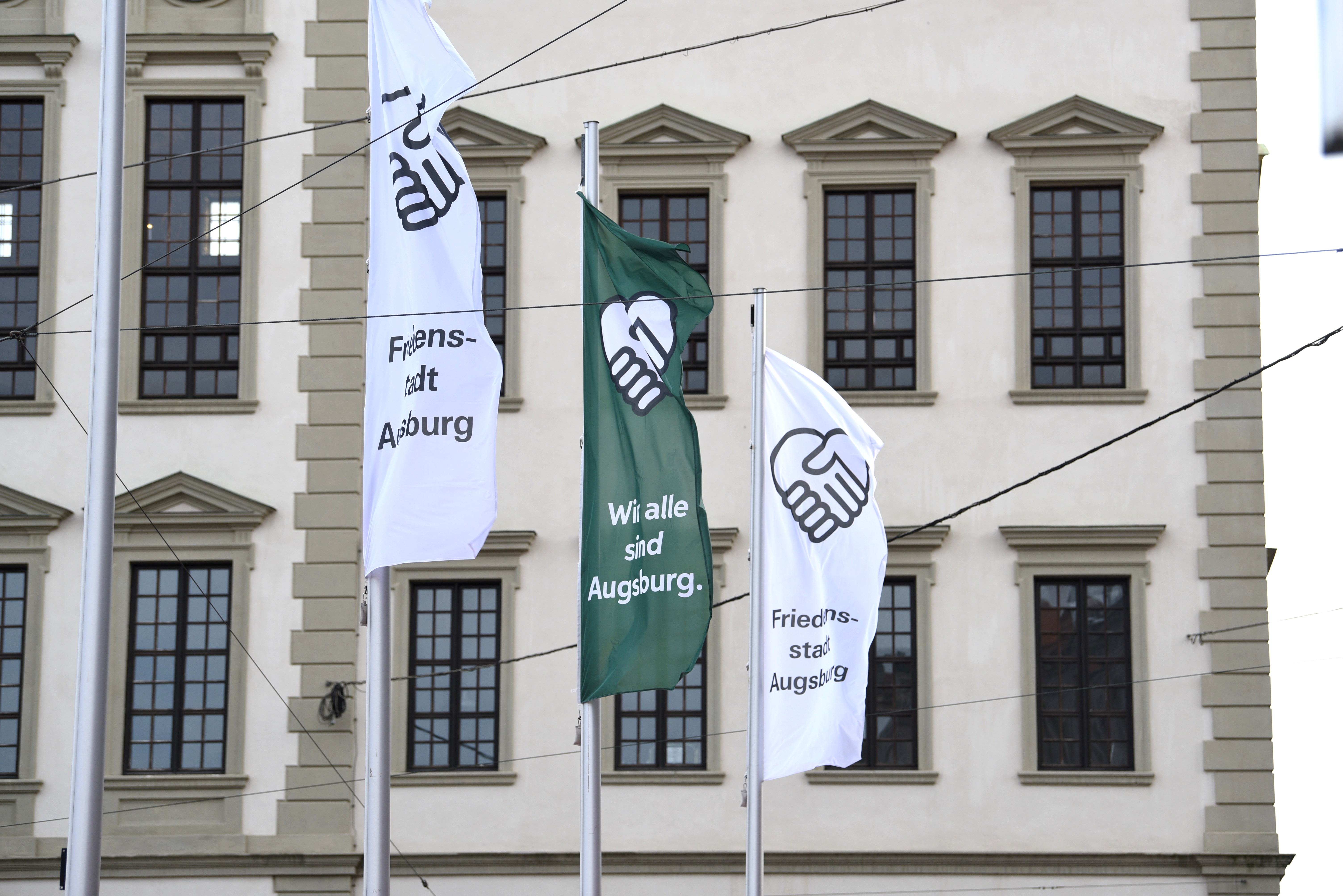 Vor dem Rathaus wehen drei Flaggen. Sie zeigen zwei Hände, die ein Herz formen. Links und rechts wehen weiße Flaggen, auf ihnen steht "Friedensstadt Augsburg". In der Mitte weht eine grüne Fahne, auf ihr steht "Wir alle sind Augsburg".