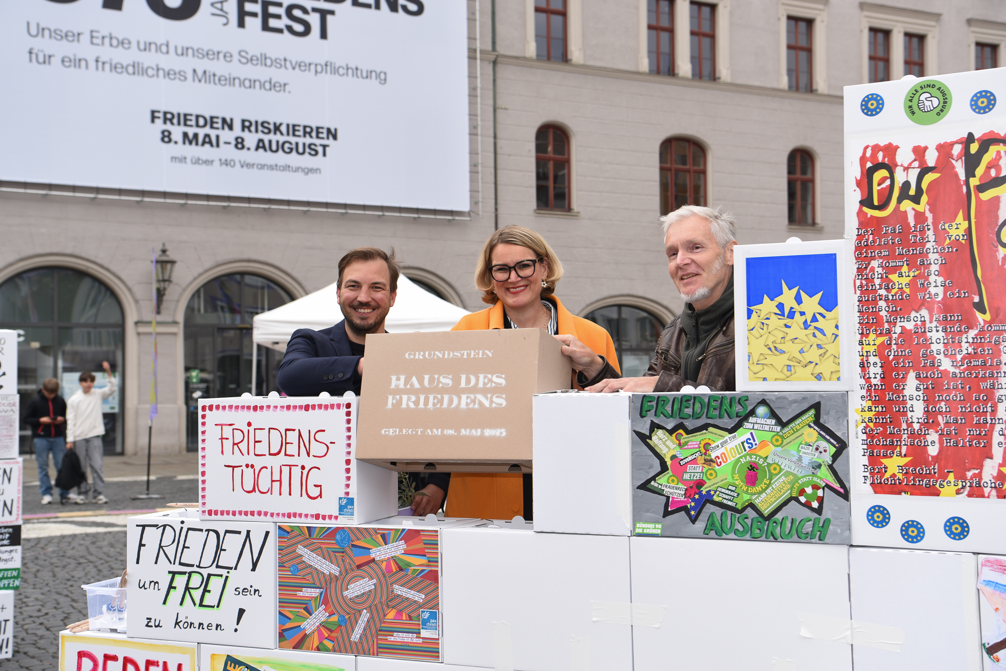 Start für ein großes Gemeinschaftsprojekt zum Friedensfest-Jubiläum. Der künstlerische Leiter des Friedensfest 2025, Eric Nikodym, OB Eva Weber und Künstler Frank Bölter stehen zwischen Pappkisten und freuen sich.